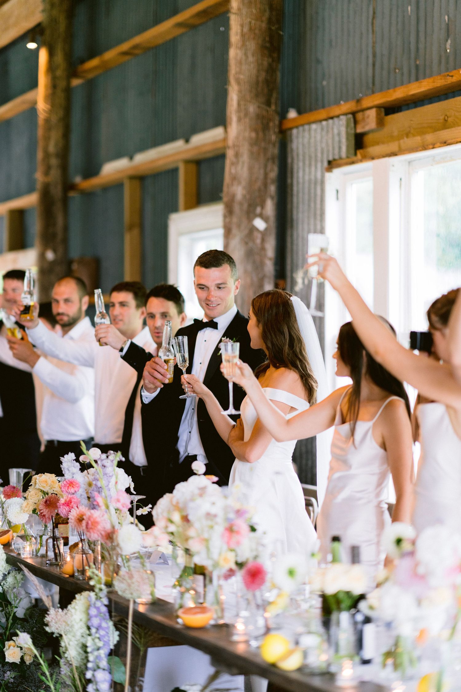 Wedding reception guests raise glasses for a toast in a rustic barn venue decorated with floral centerpieces.