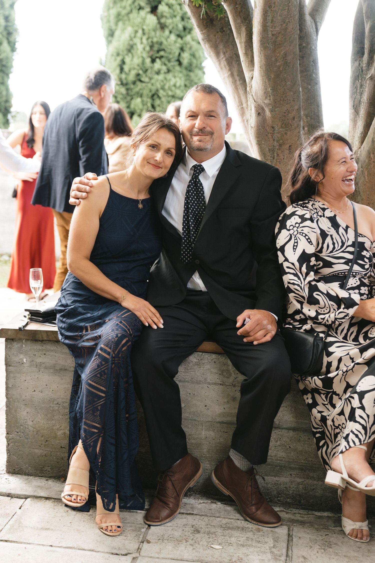 Wedding guests sit casually on a stone bench sharing candid moments and laughter.