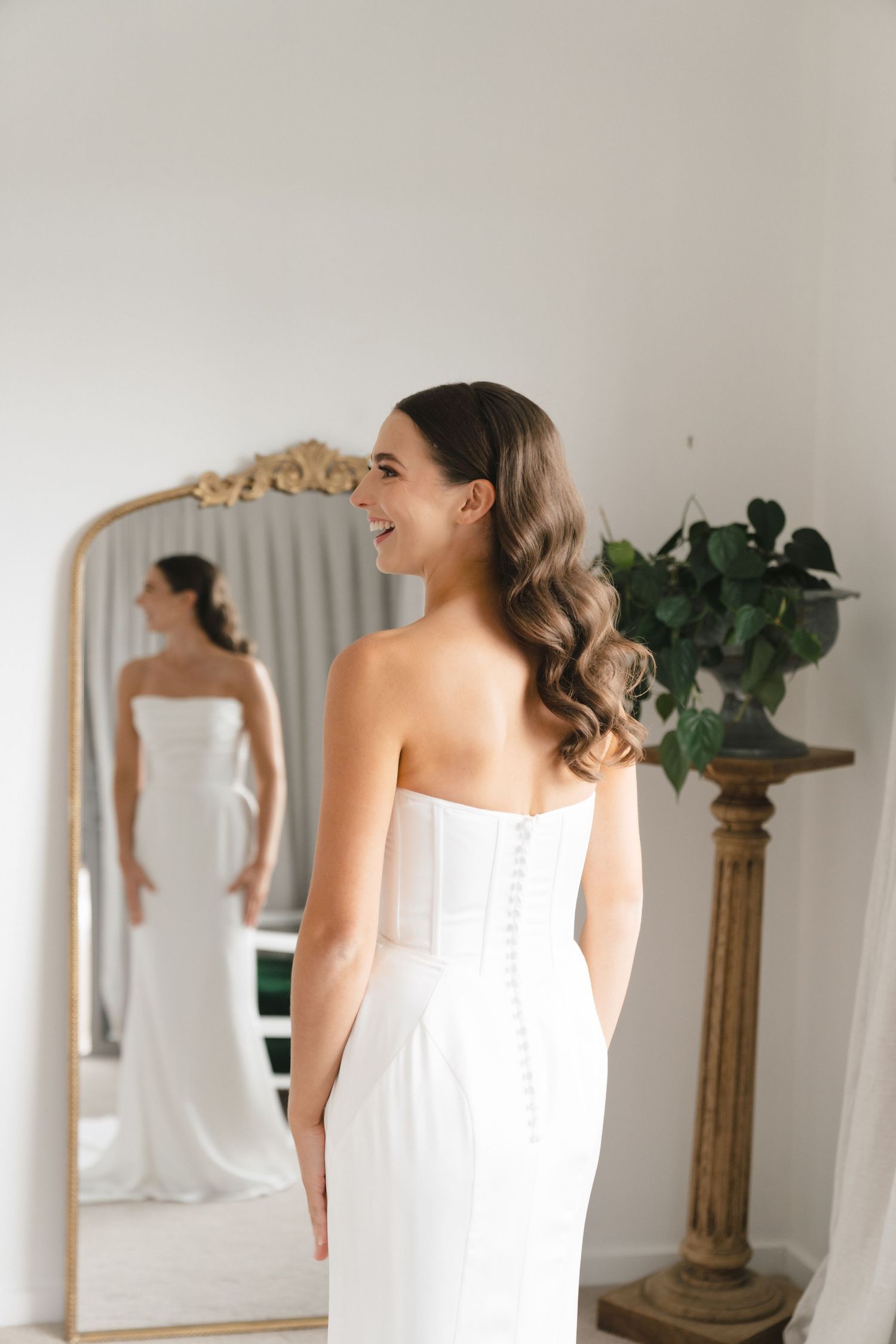 Bride in white wedding dress looking at reflection in ornate mirror.