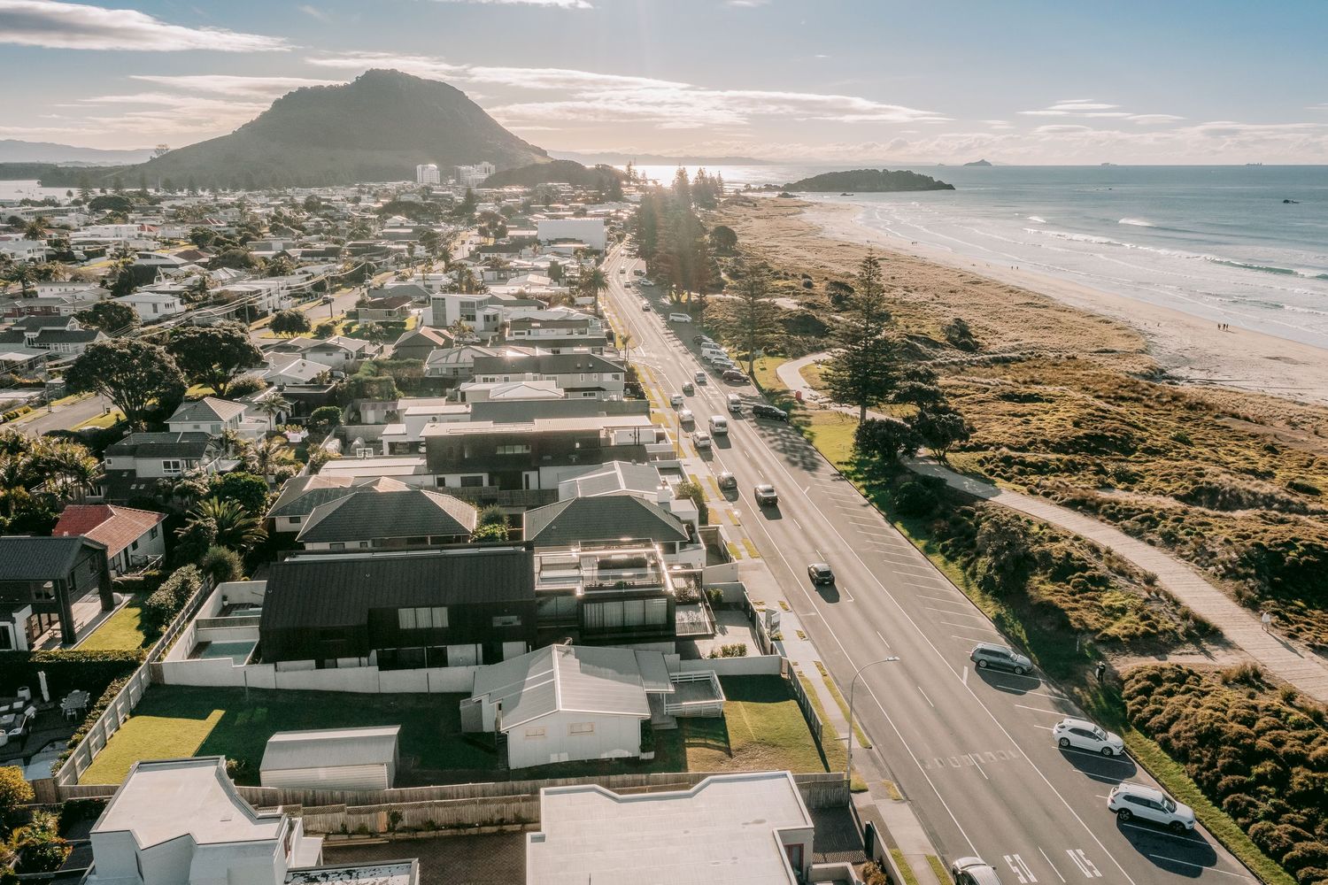 Coastal town with beachfront homes and a scenic mountain backdrop along a winding shoreline road.