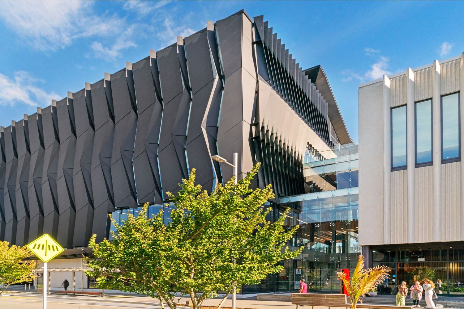 Modern university building with angular architectural features and yellow tree against blue sky.