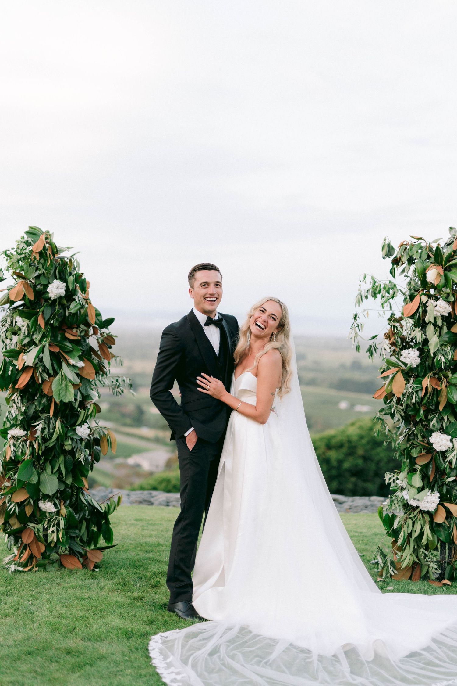 A couple poses in an elegant outdoor wedding setting with greenery-adorned arches on rolling hills.