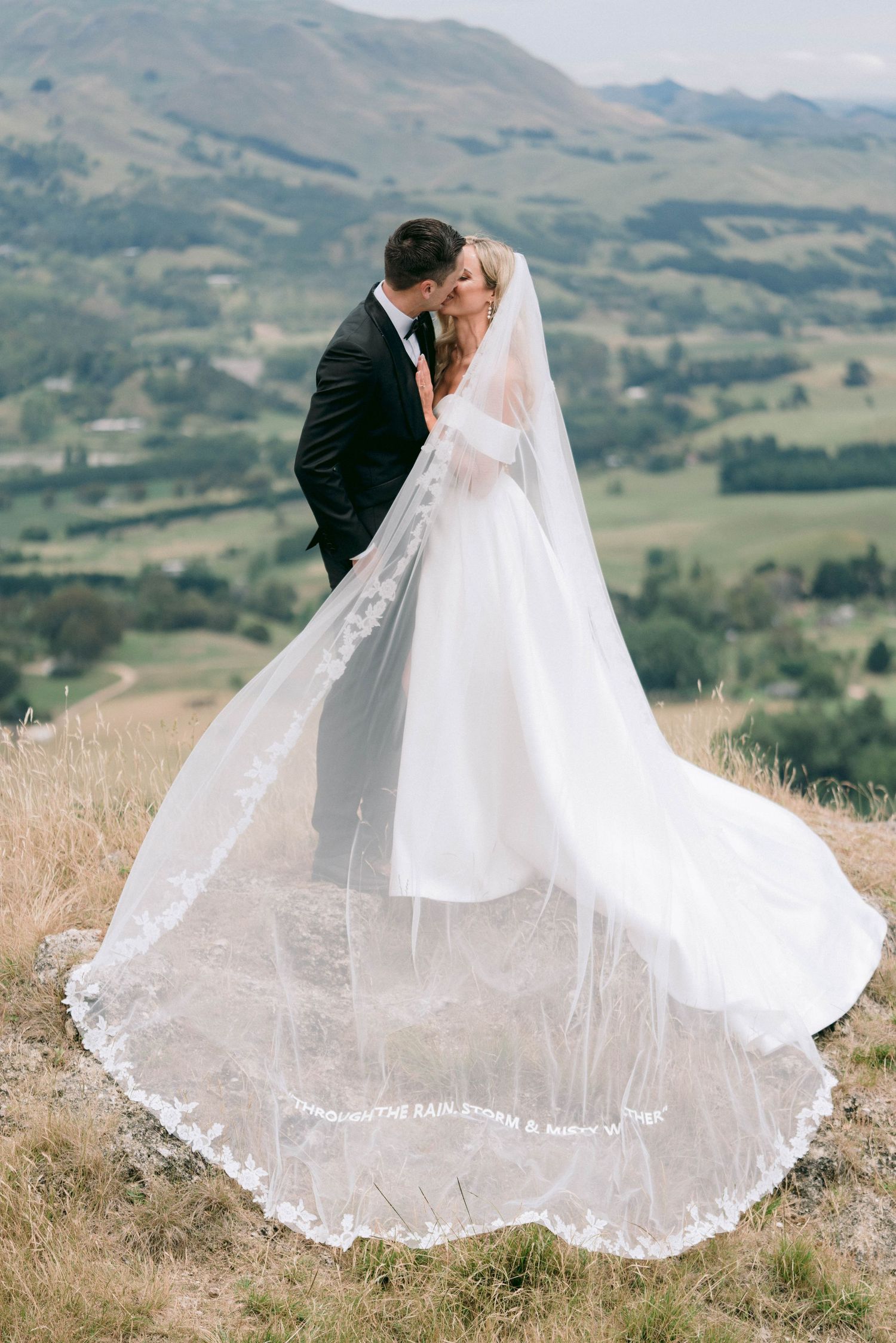 Romantic wedding portrait on a hillside with flowing white veil and mountain backdrop.