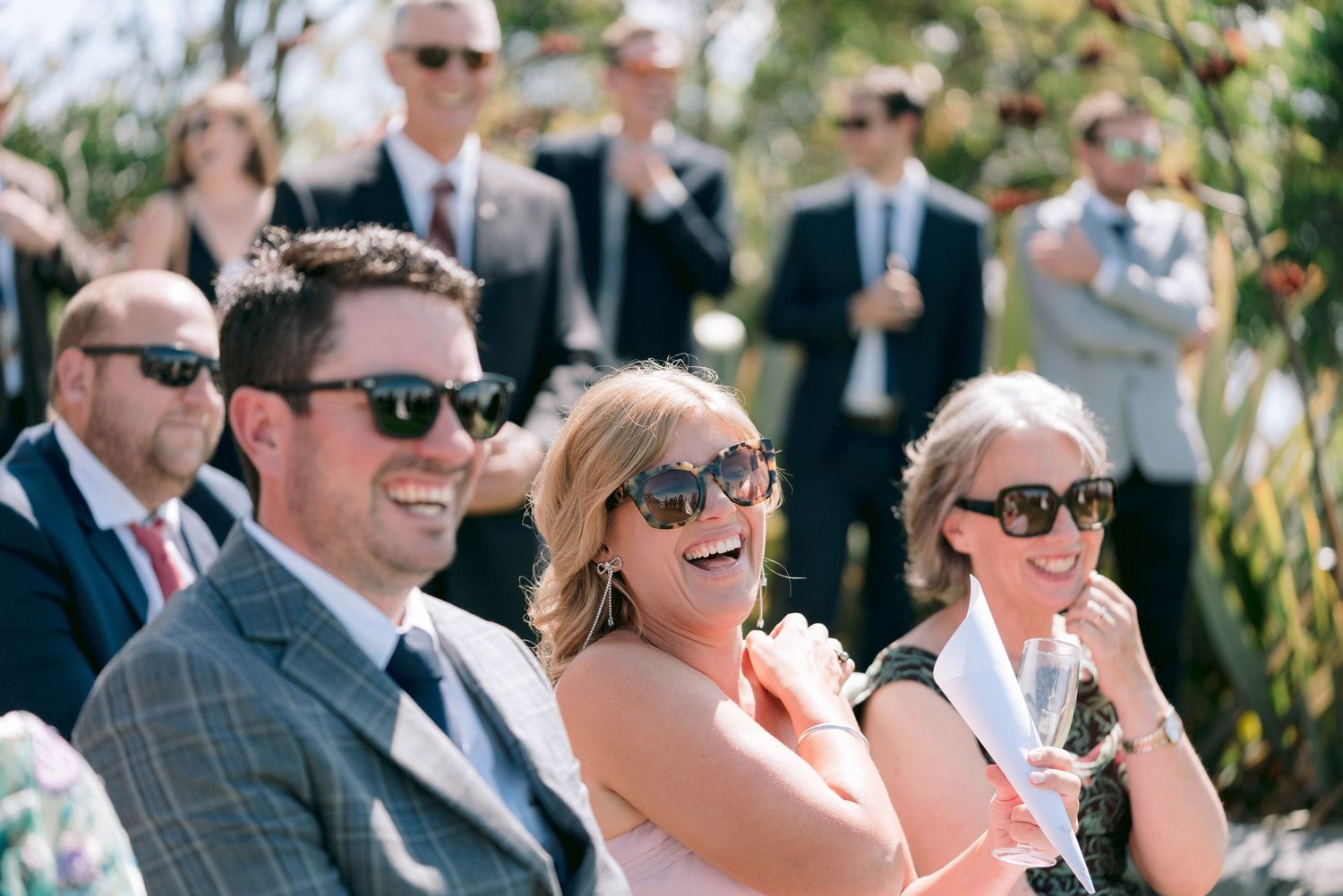 Wedding guests in sunglasses laugh joyfully while seated at an outdoor ceremony.