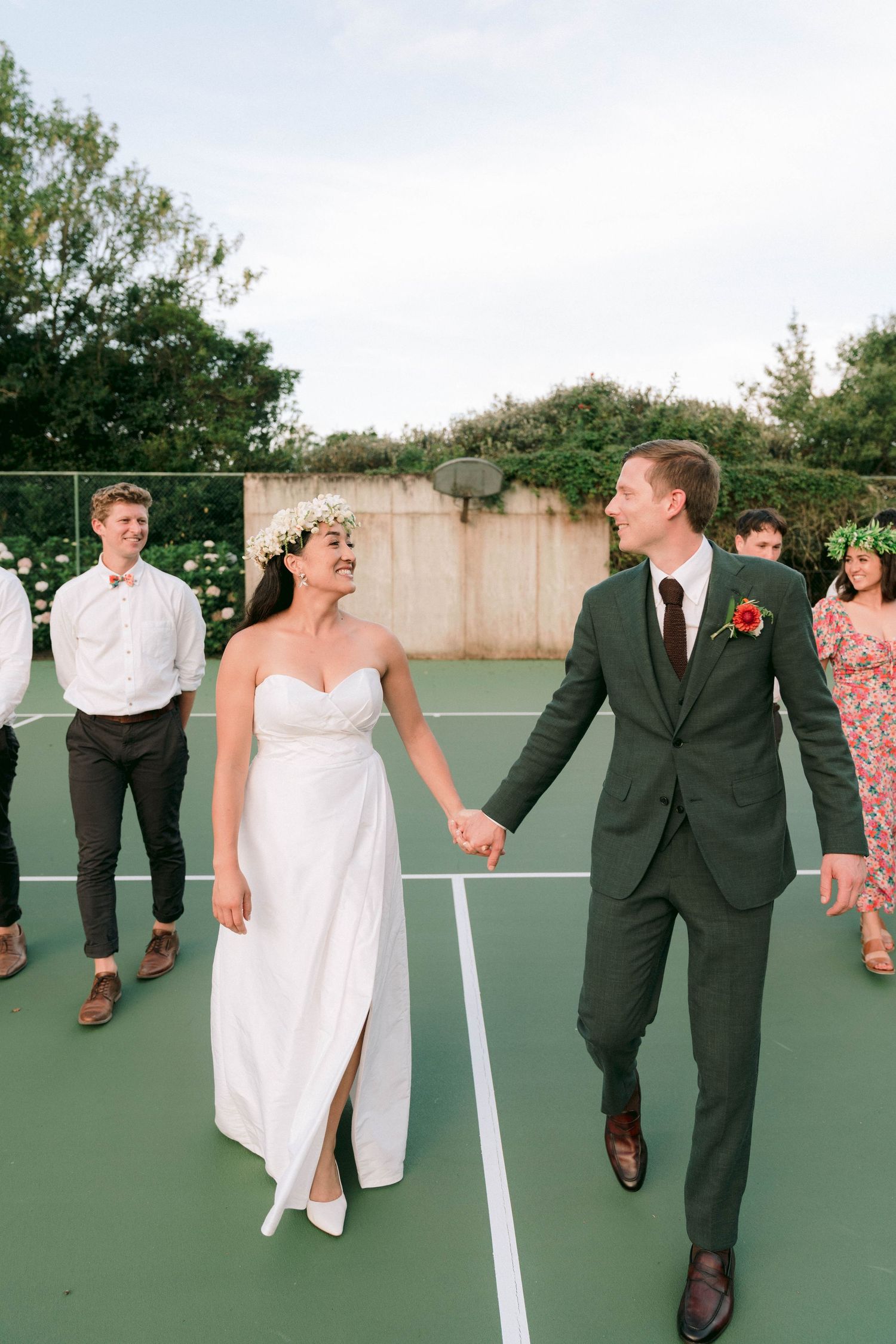 A newlywed couple walks hand in hand on a green tennis court with guests looking on.