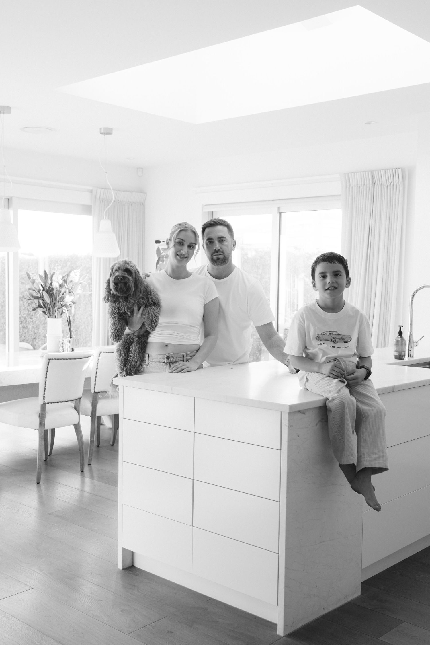 A family gathered around a white kitchen island with their pet dog in a bright sunlit room.