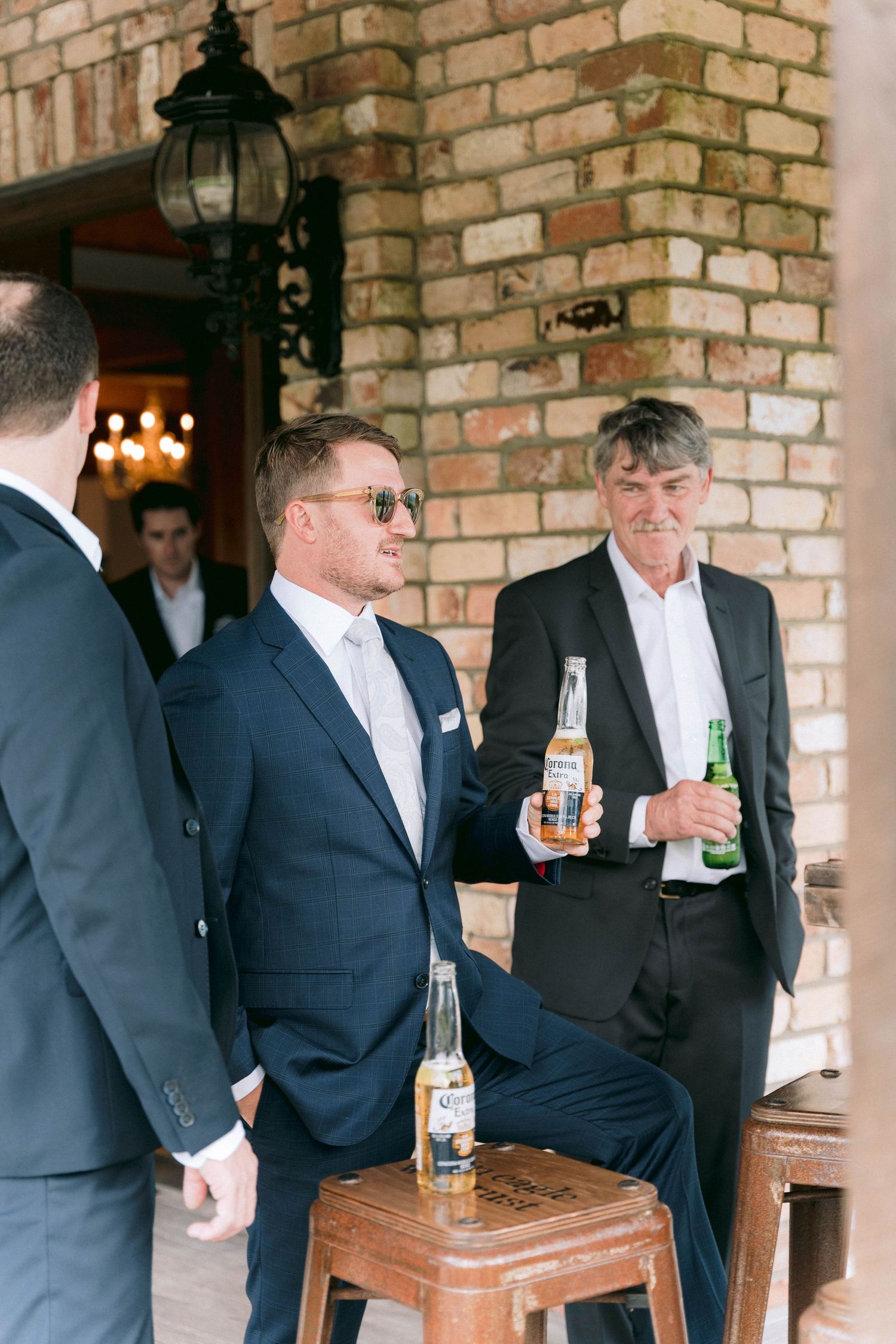 People in suits holding beer bottles gather near a brick wall with a classic outdoor lantern during what appears to be an event.