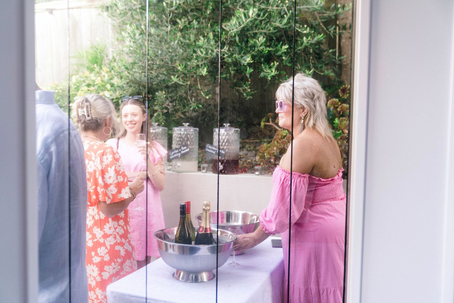View through window of party guests gathered around drinks and refreshments outdoors.
