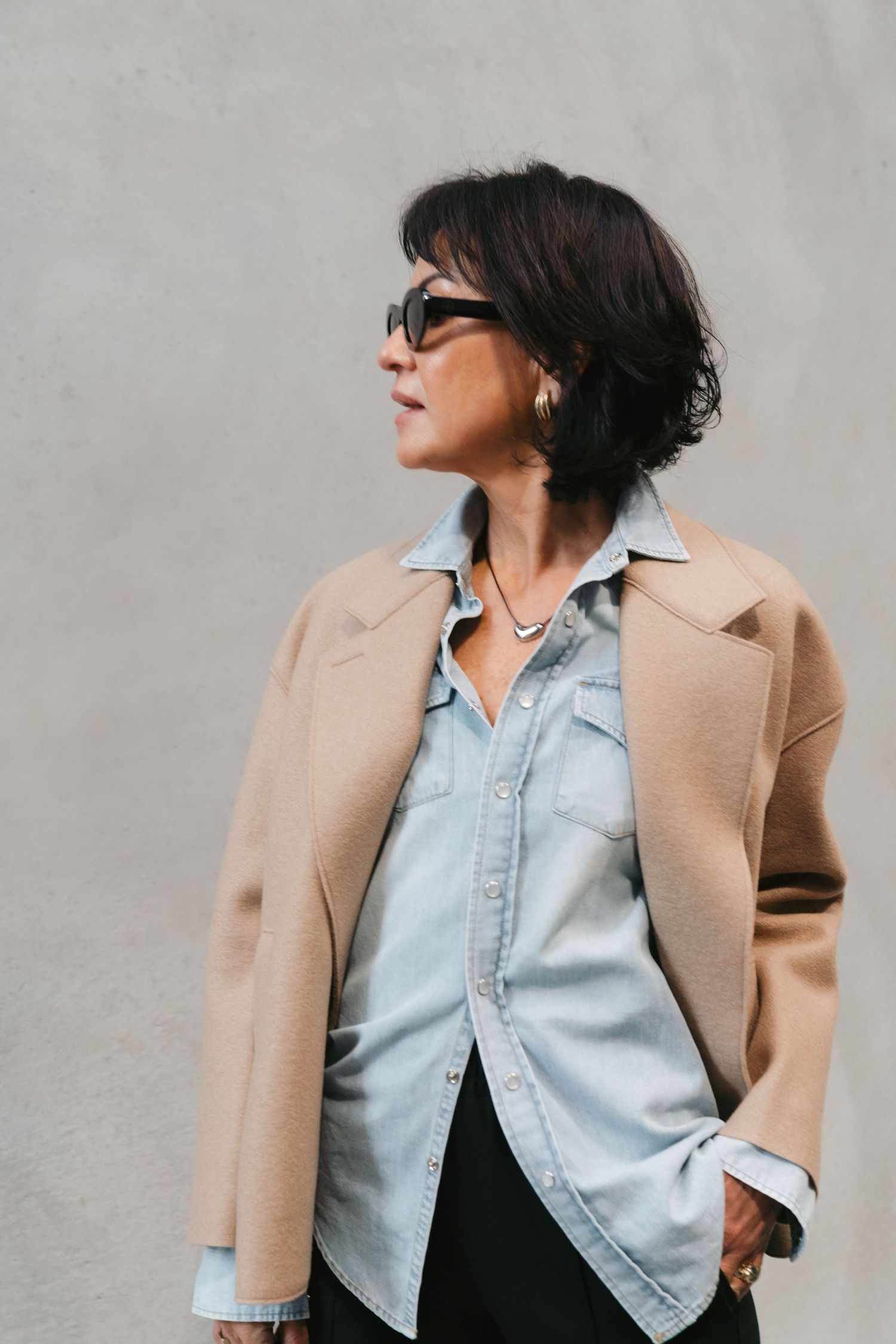 Stylish layered winter outfit with denim shirt, camel coat, black sunglasses and casual short dark hair.