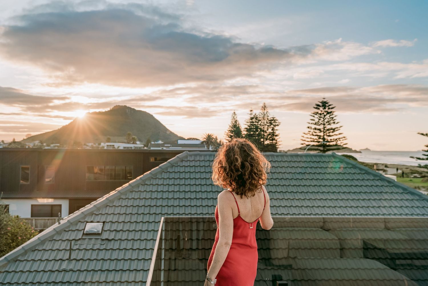 Person in red dress watching sunset over coastal town with mountain silhouette in background.