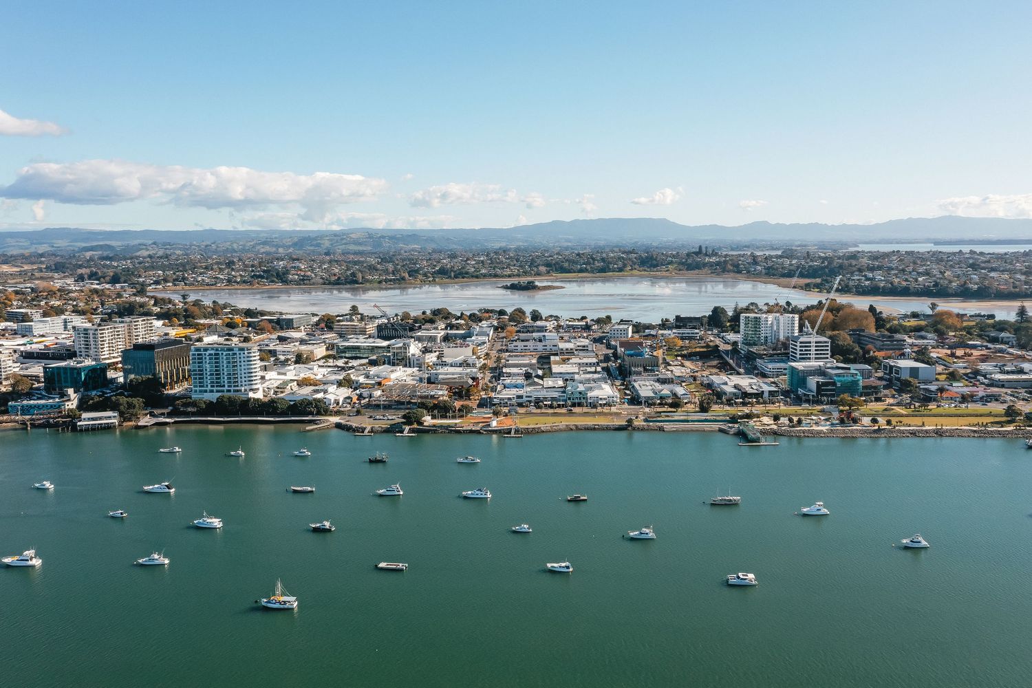 A panoramic aerial view of a seaside harbor with boats moored in the blue waters and coastal buildings along the shore.