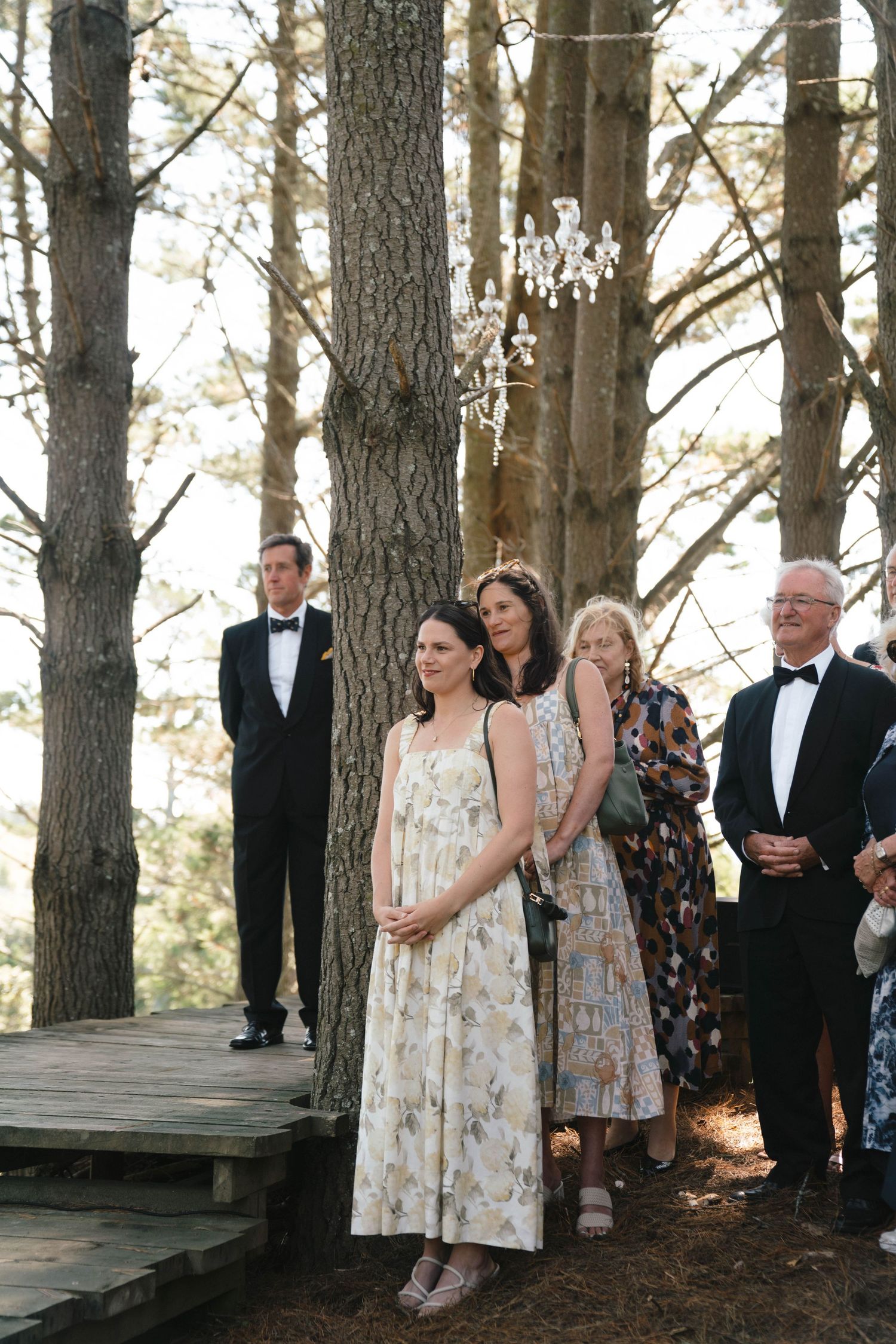 Wedding guests in formal attire standing on a wooden platform among tall pine trees.