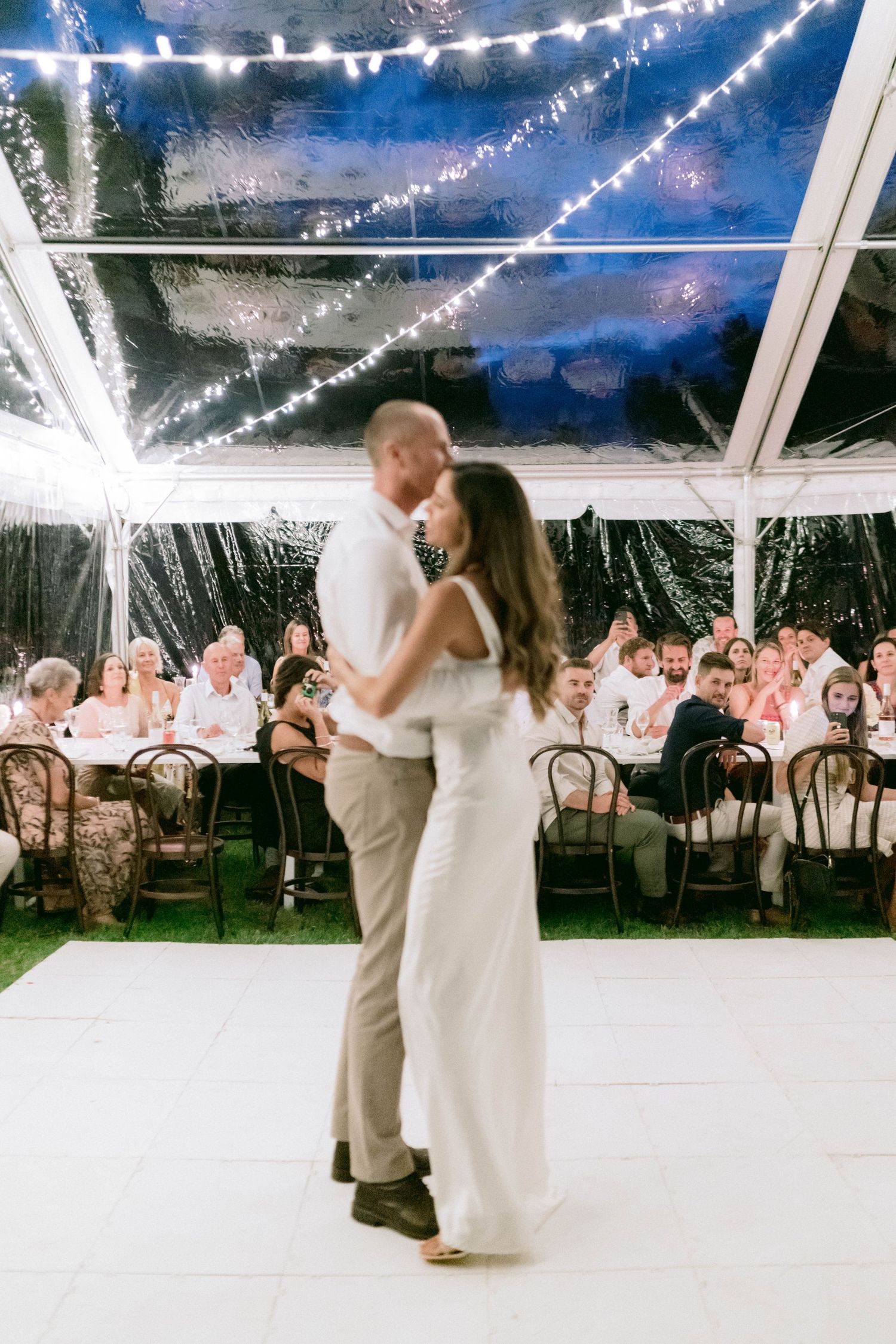 A couple shares their first dance under twinkling lights in a clear-top tent surrounded by guests.