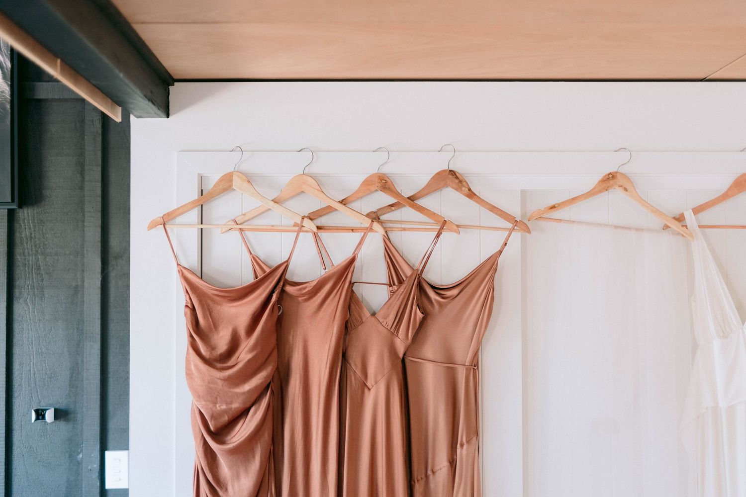 Neutral champagne-colored bridesmaid dresses hanging on wooden hangers against a white wall.