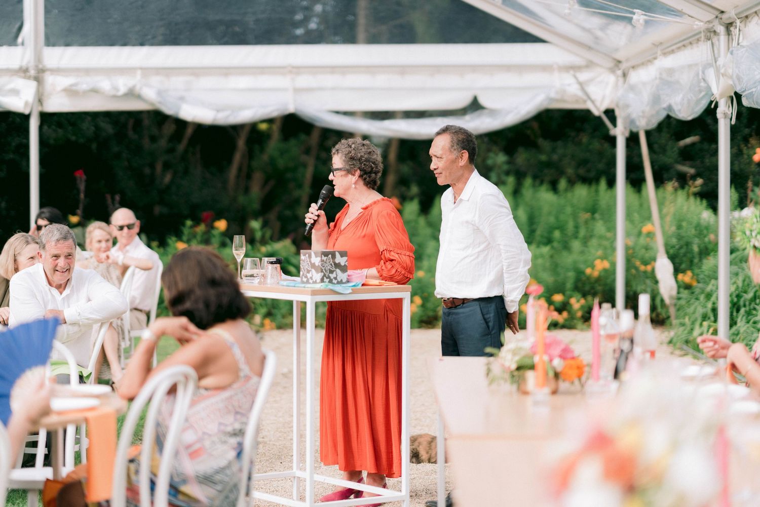 People gathered under a white tent for speeches at an outdoor reception.