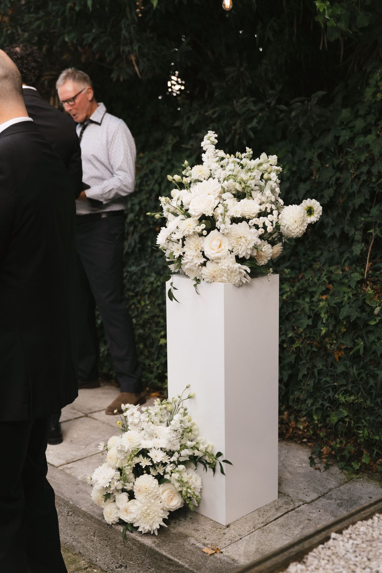 Elegant white floral arrangement displayed on tall pedestal at outdoor event.