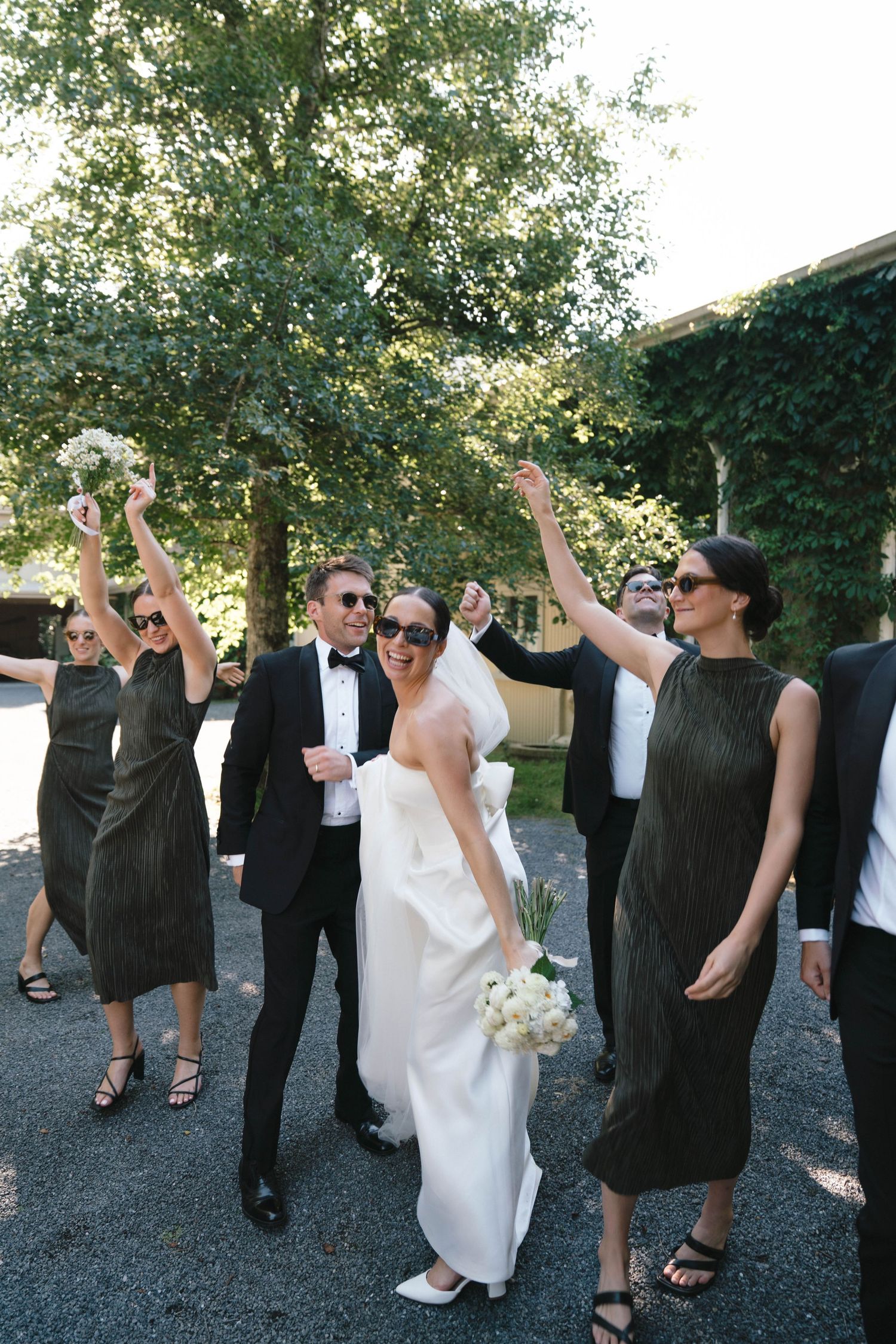 Wedding party celebrates outdoors with arms raised in joy while bride holds white bouquet and wears elegant gown.