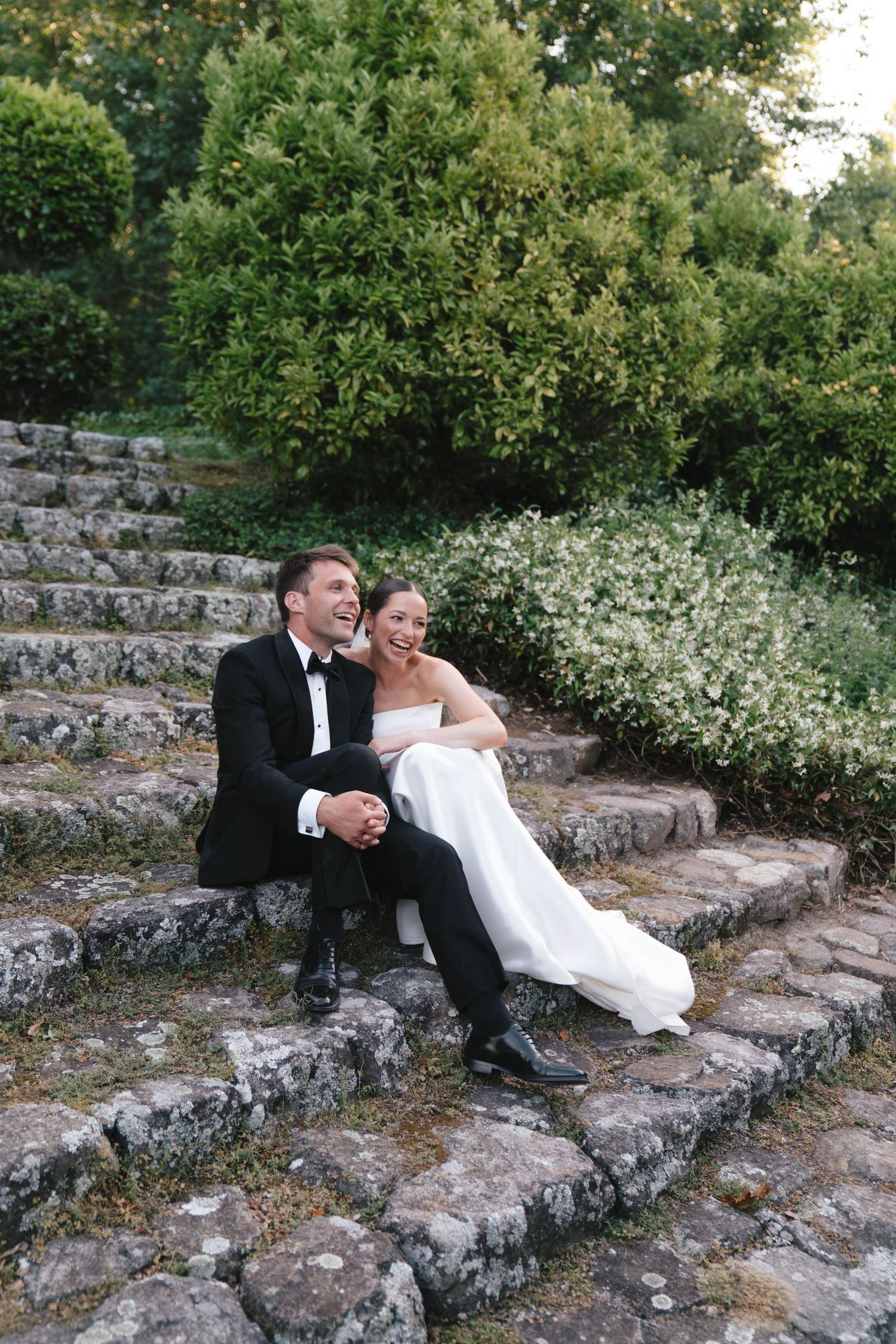 A couple in wedding attire sits together on stone steps surrounded by lush greenery.