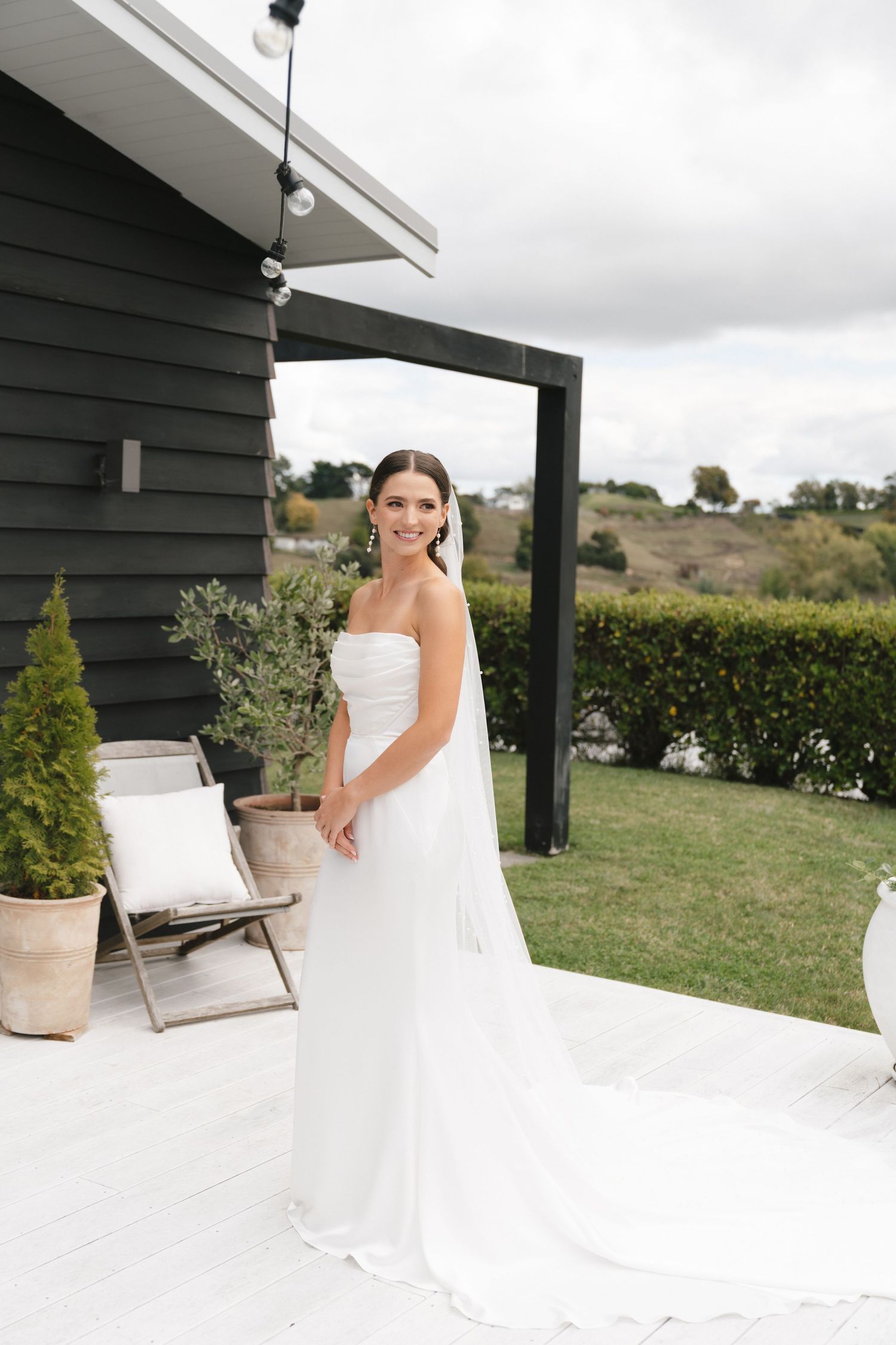 Person in elegant white wedding gown posing on patio with scenic countryside view.