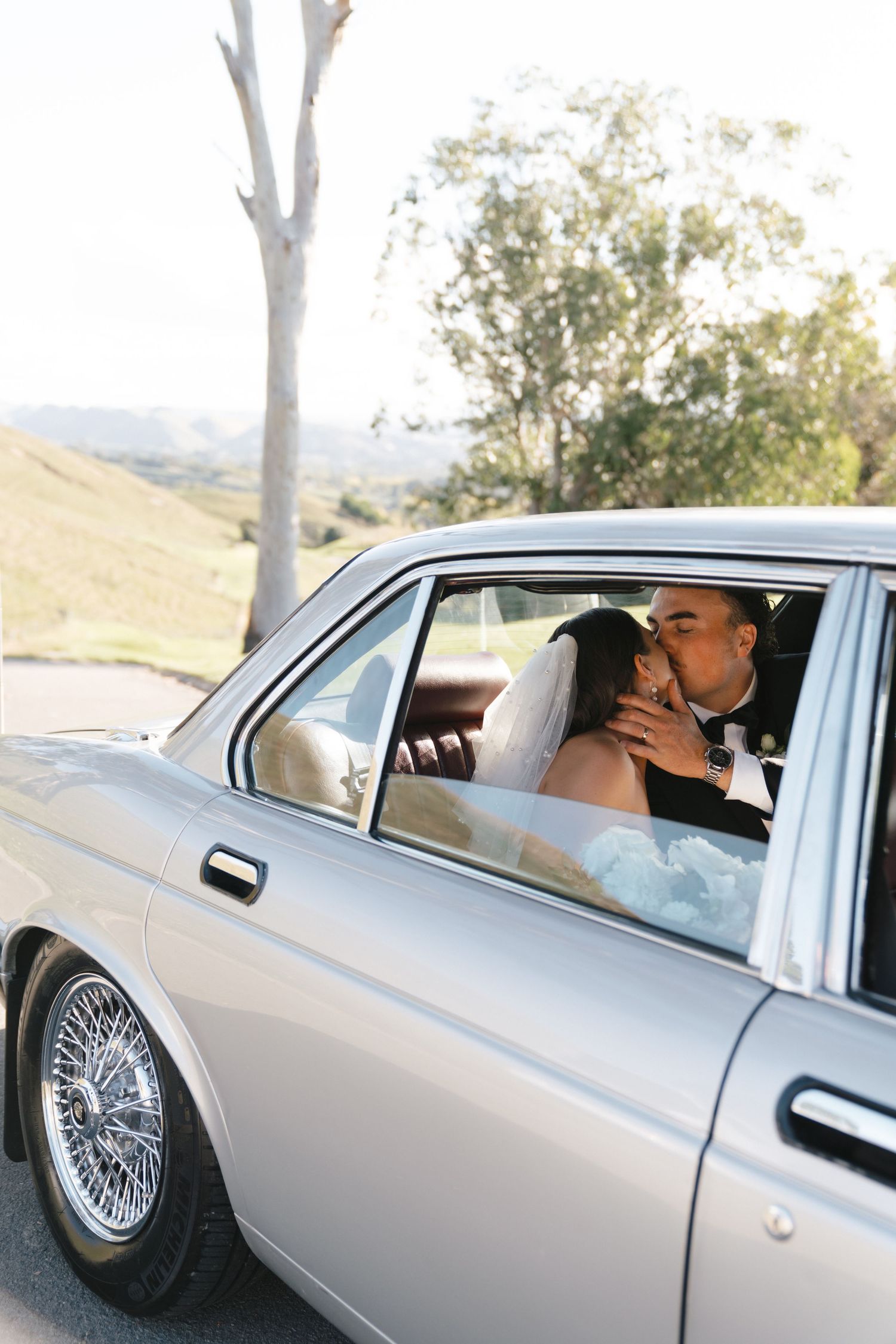 Three views of a luxurious silver sedan with classic wire-spoke wheels parked outdoors against a hilly landscape.