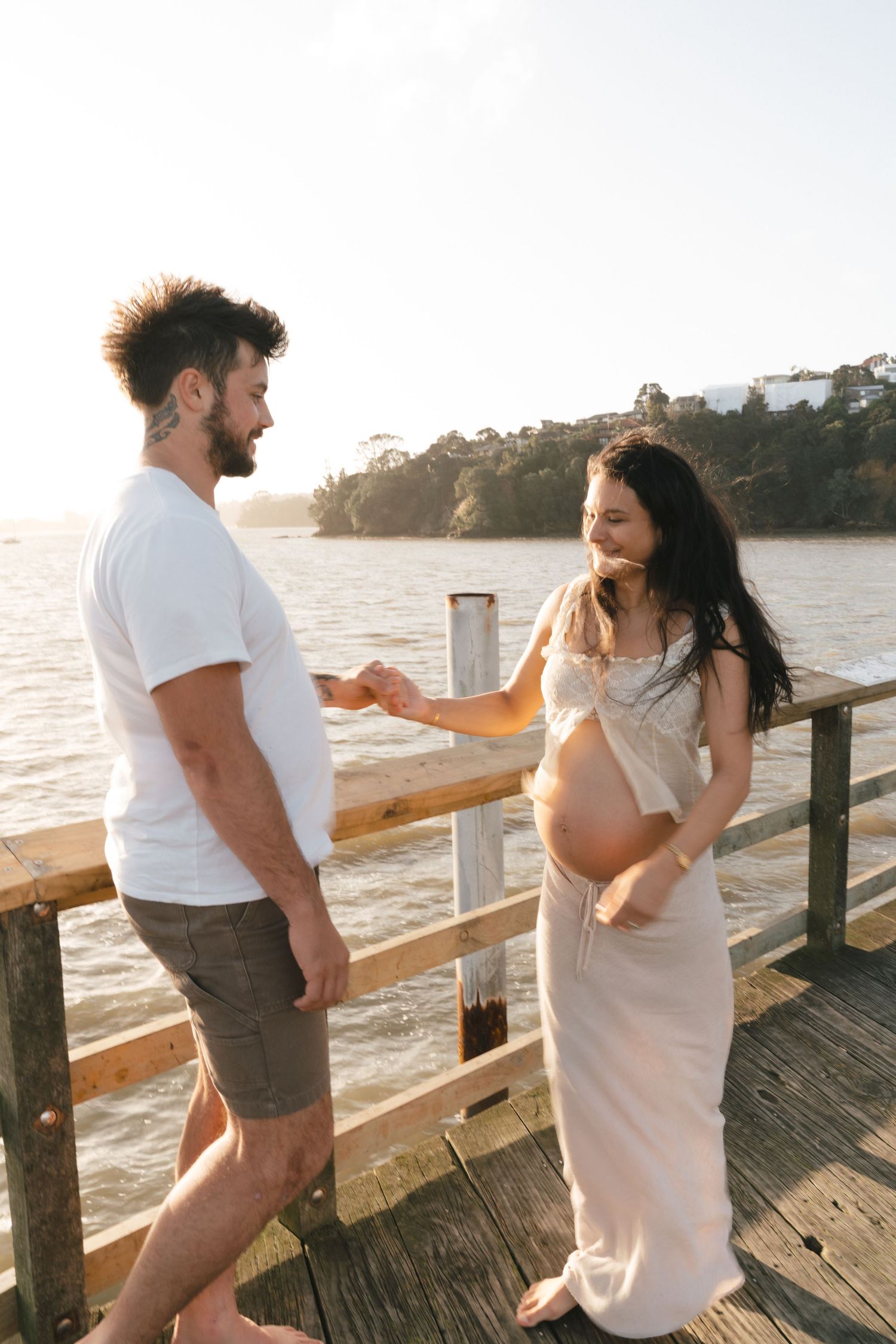 Couple sharing a tender moment on a wooden pier at sunset with the pregnant woman wearing a flowing white dress.