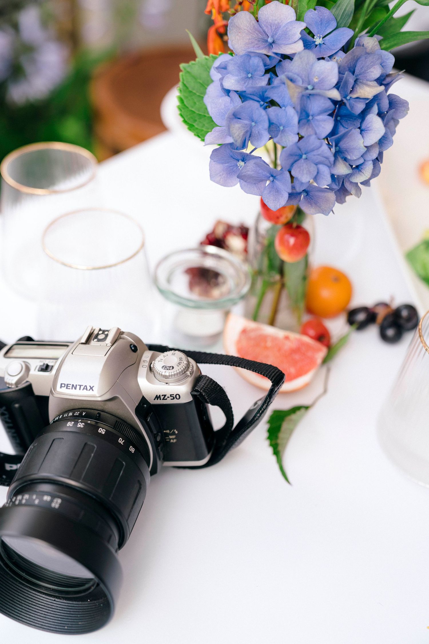 A silver camera sits on a white table with blue hydrangeas, fruits, and glasses in the background.