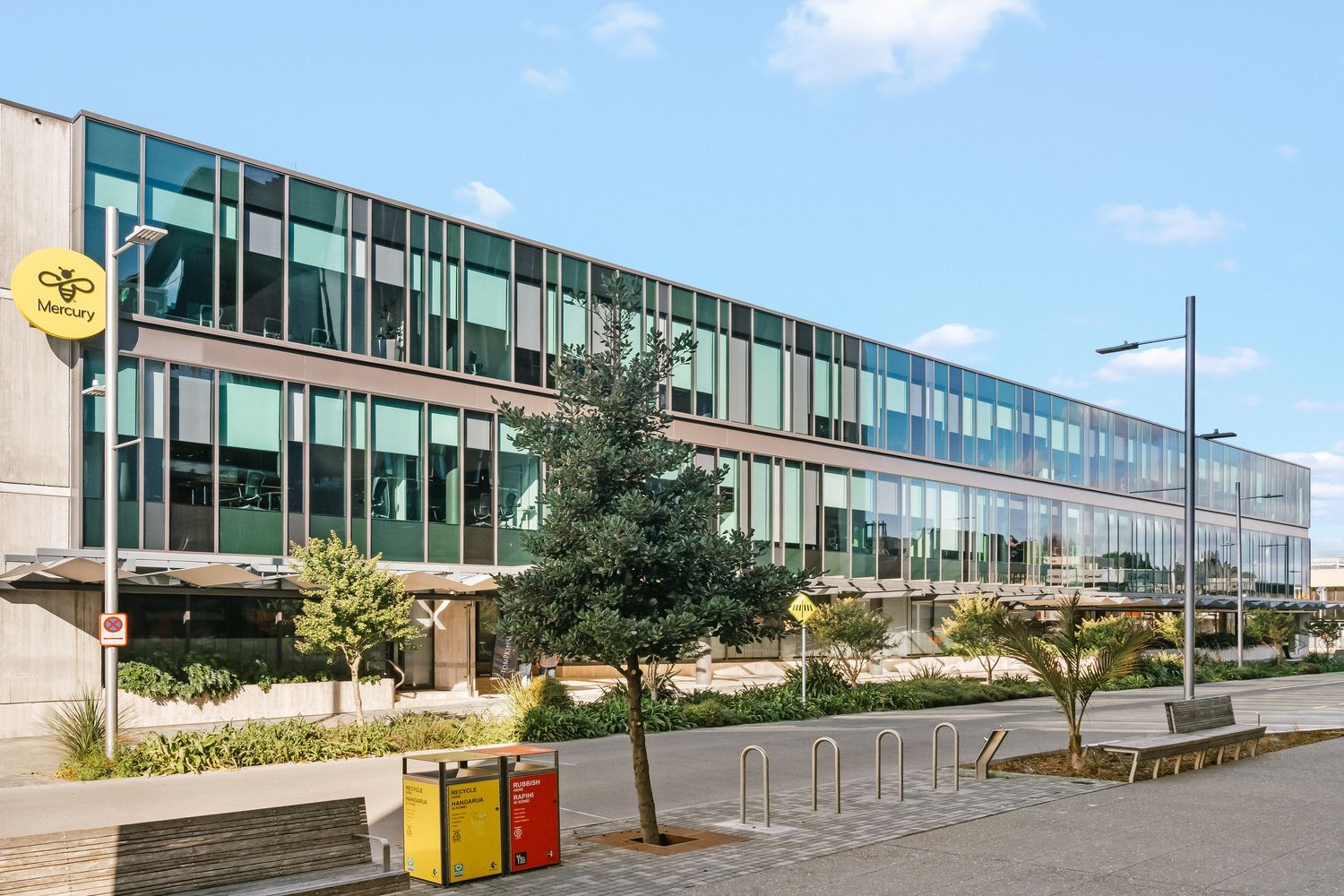 Modern office building exterior with glass facade and landscaped walkway along a sunny parking area.