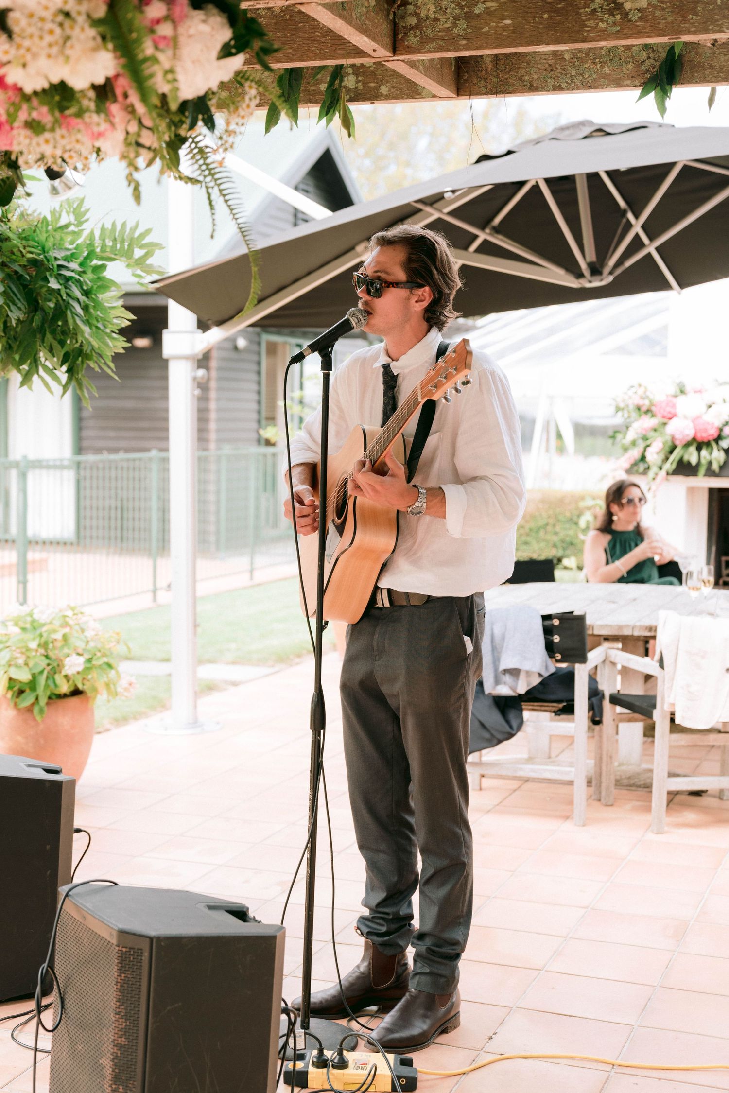A musician performs with an acoustic guitar under a white outdoor tent with floral decorations.