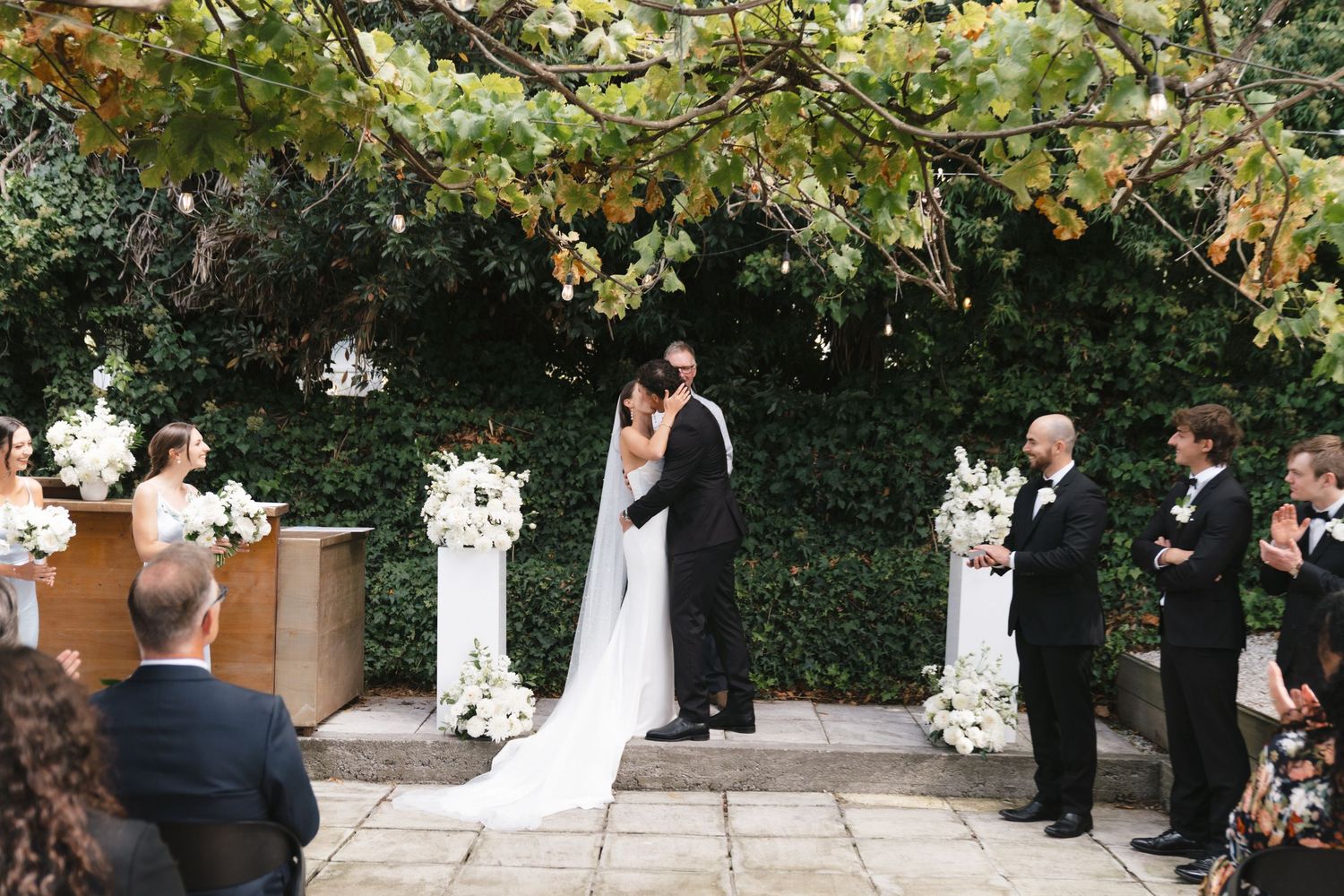 Wedding party gathered in a lush garden setting with white floral arrangements.