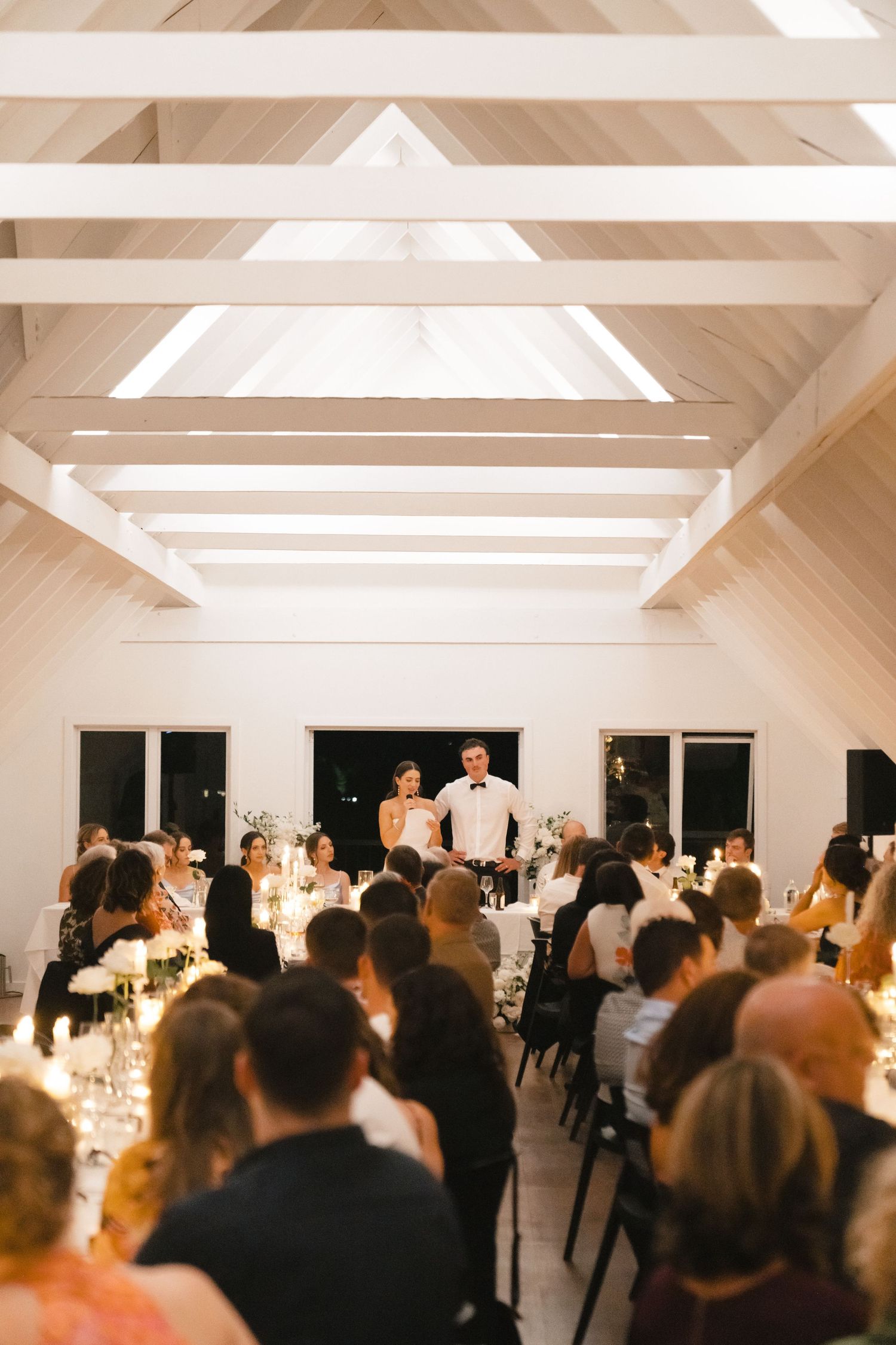 White vaulted ceiling reception venue with guests gathered at elegant tables for a wedding celebration.