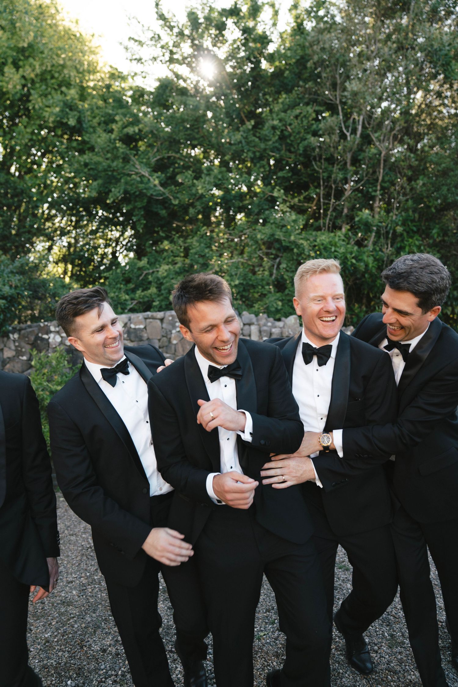 A group of groomsmen in black tuxedos and bow ties laugh together outdoors during a wedding photo session.