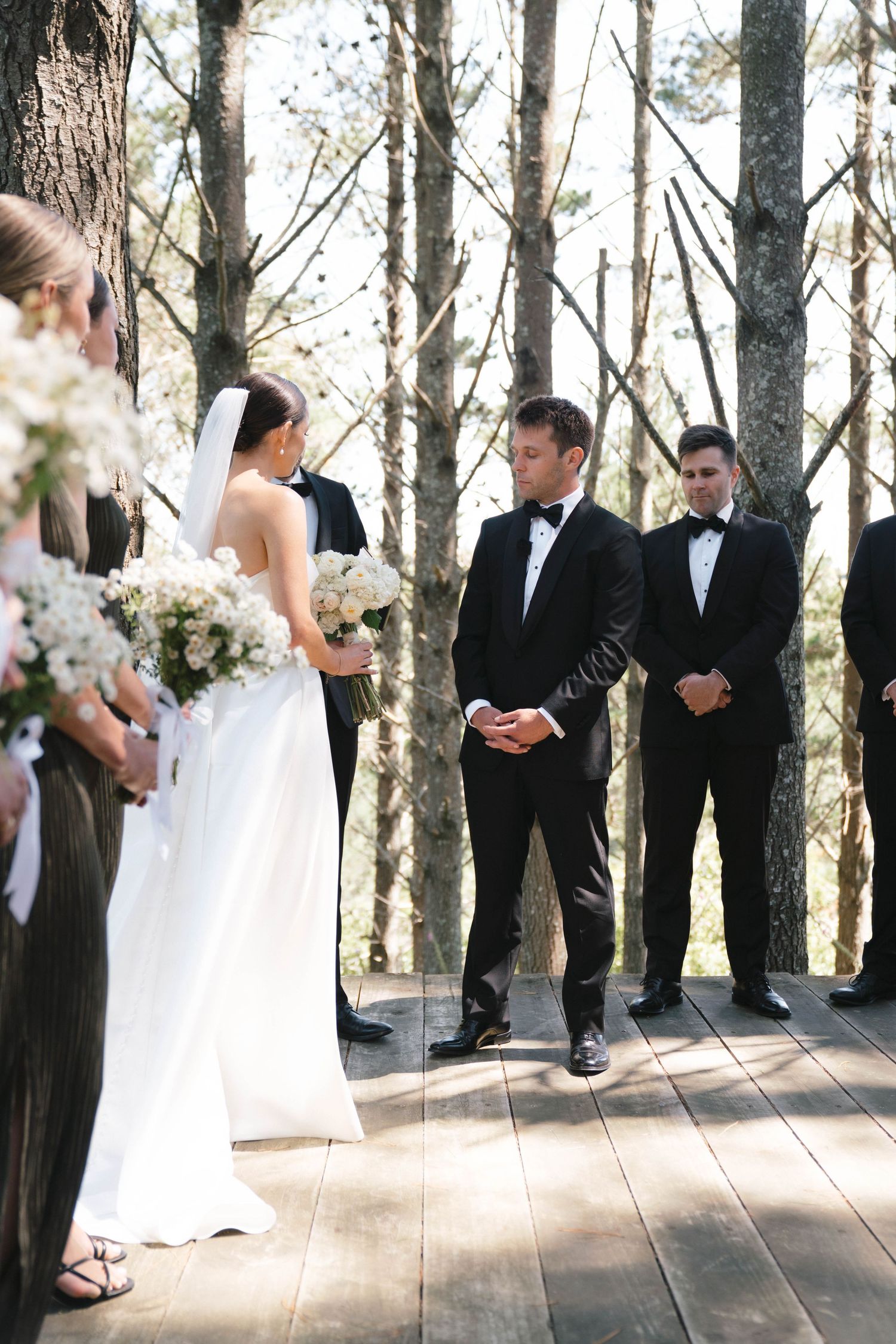 An elegant outdoor wedding ceremony takes place on a wooden deck surrounded by tall trees in a forest setting.