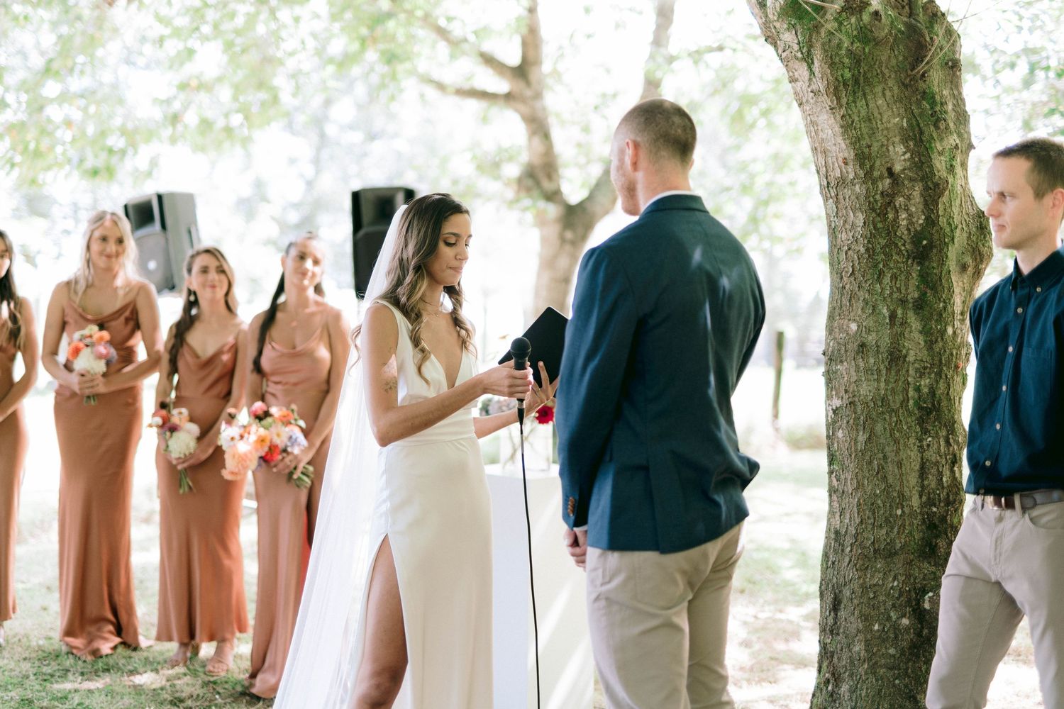 Outdoor wedding ceremony under trees with bridesmaids in coral dresses watching the couple exchange vows.