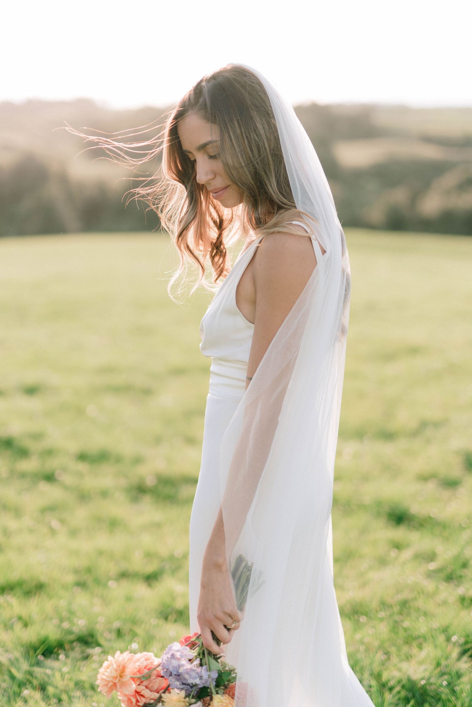 Delicate floral bouquet with coral and purple blooms held against a sunlit field with soft dreamy lighting.