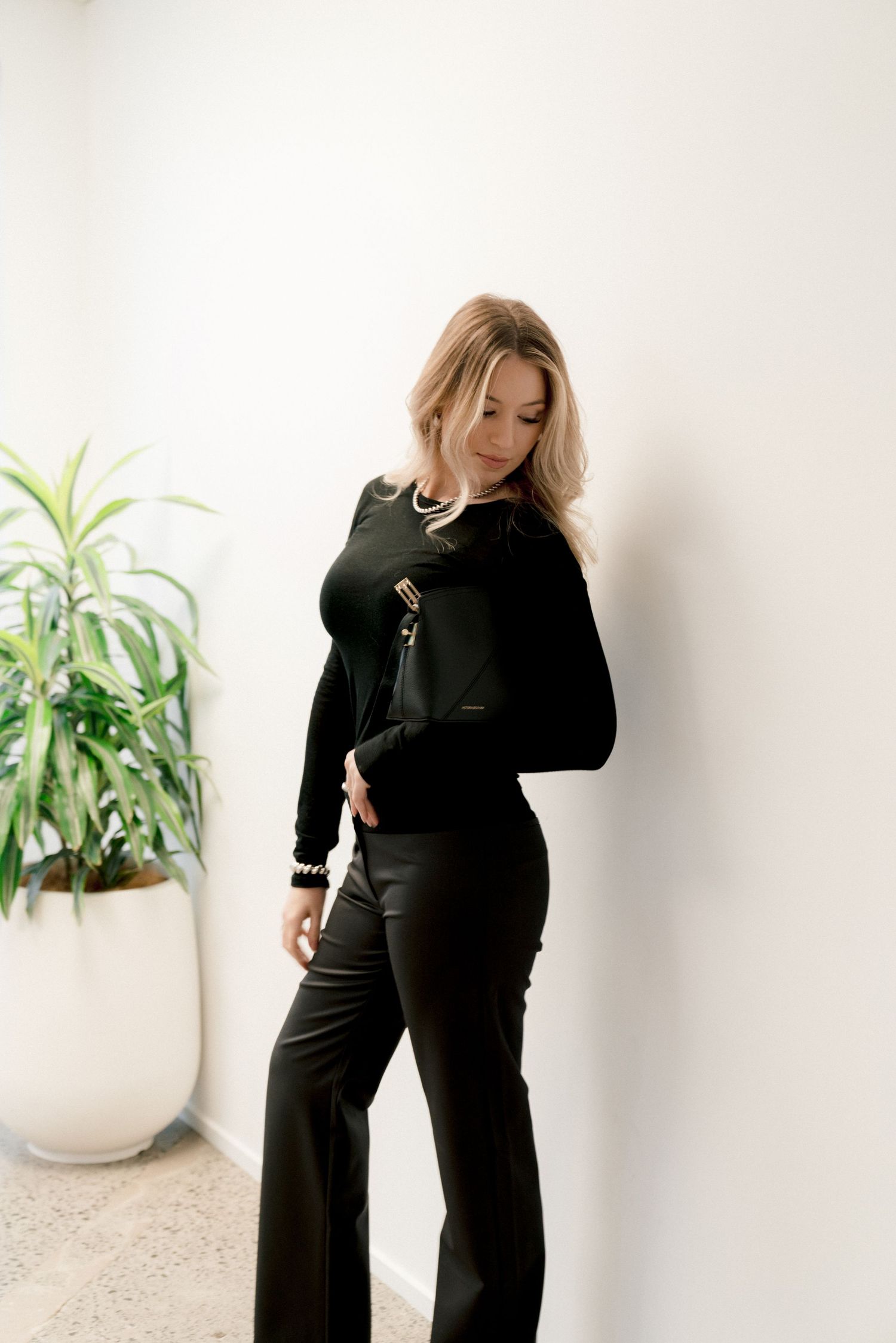 Elegant all-black outfit with flared pants and long sleeves photographed against white wall with potted plant decor.