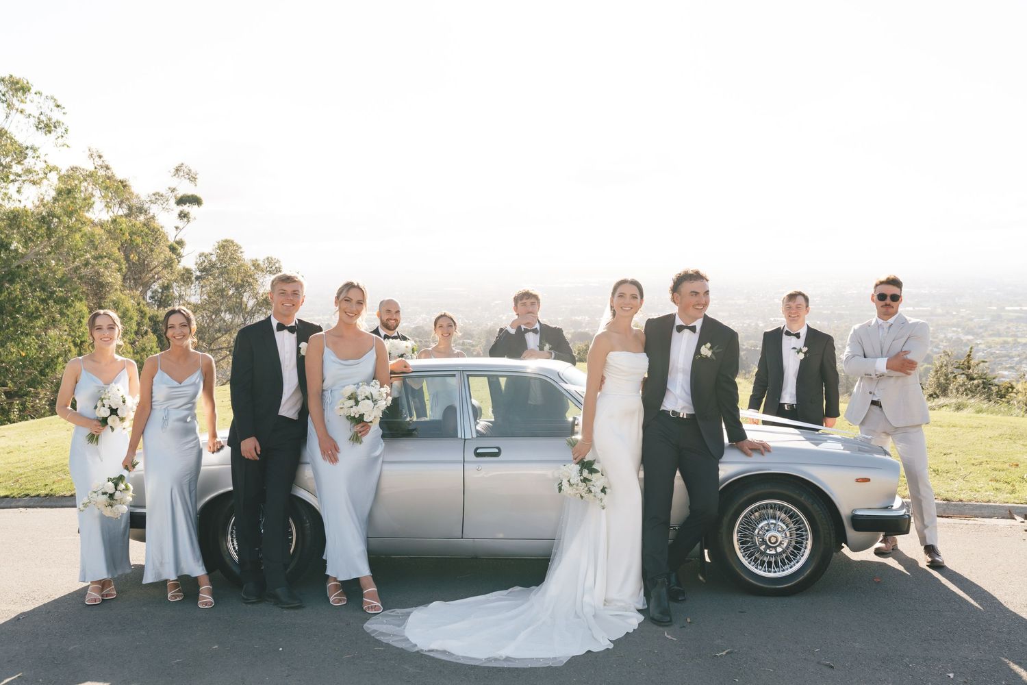 Wedding party poses with classic silver car outdoors on sunny day.