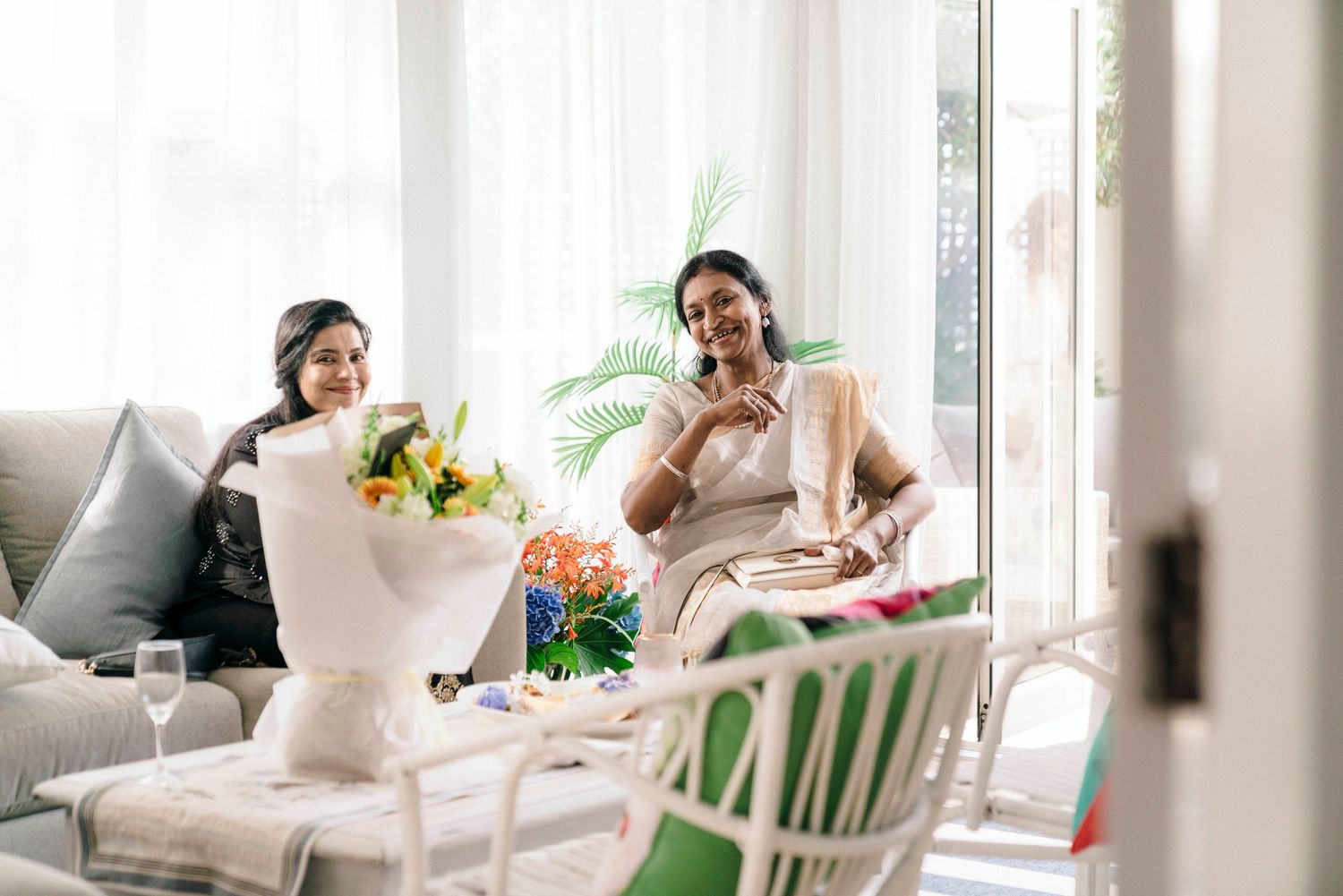 Two people relaxing on a light gray couch with flowers and greenery in a bright modern living room setting.