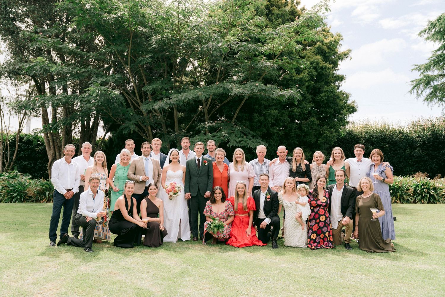 Large wedding group photo with guests posed outdoors under trees.
