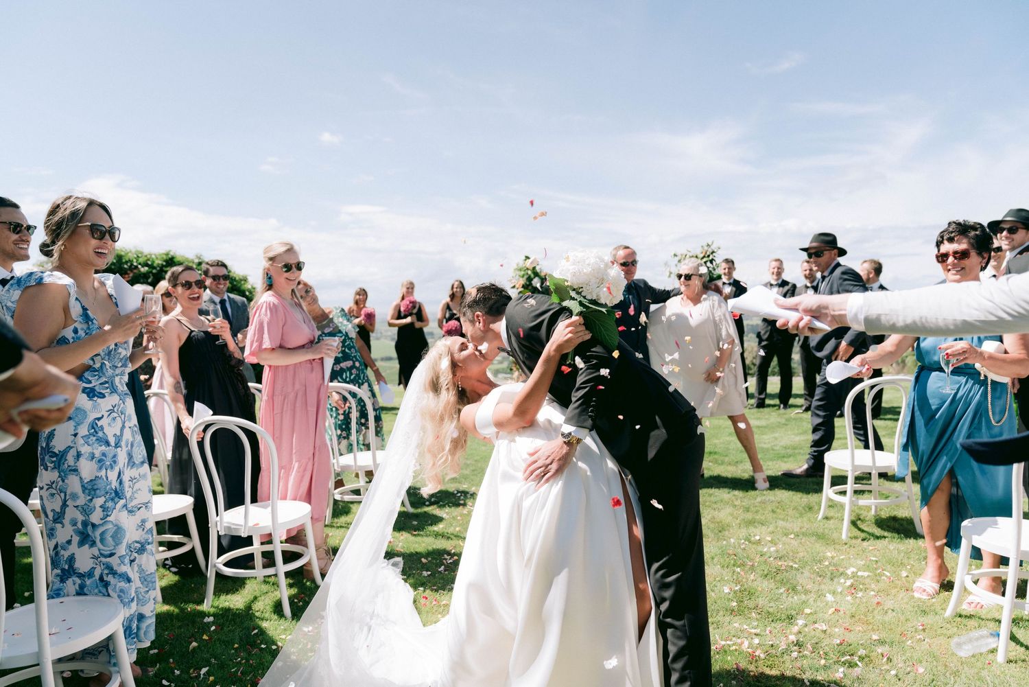 Wedding guests gather outdoors on a grassy hilltop during a sunny celebration.