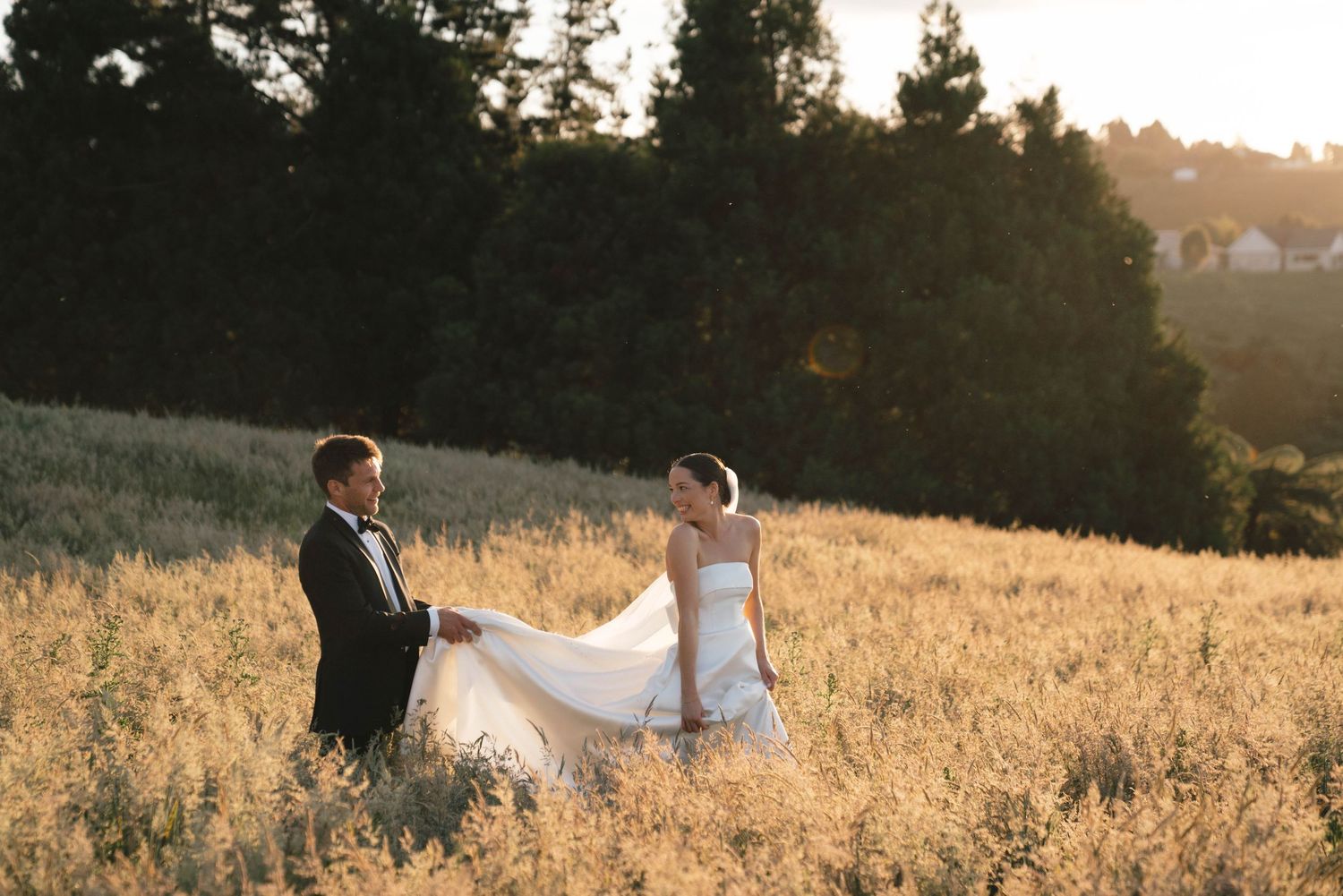 Two people in wedding attire stand together in a golden sunlit meadow at sunset with pine trees in the background.