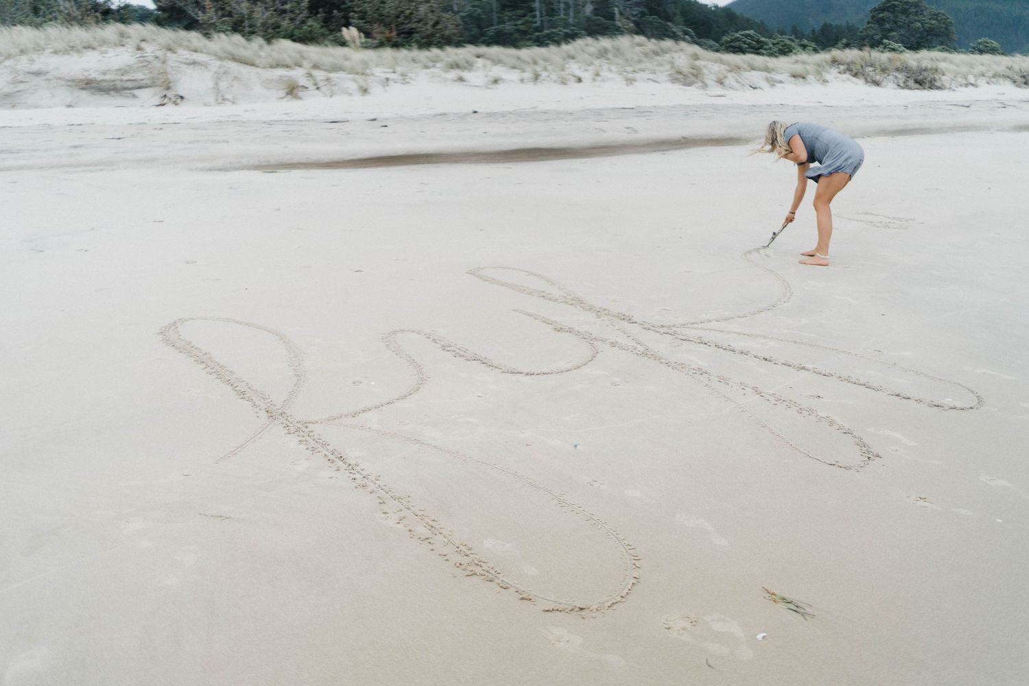 Person writing in sand on empty beach with mountains in background.
