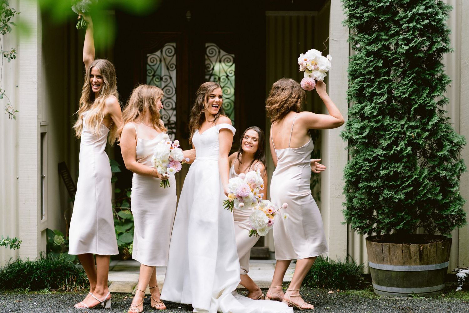 Bridesmaids in white dresses gather outdoors in front of an ivy-covered wall at an elegant venue.