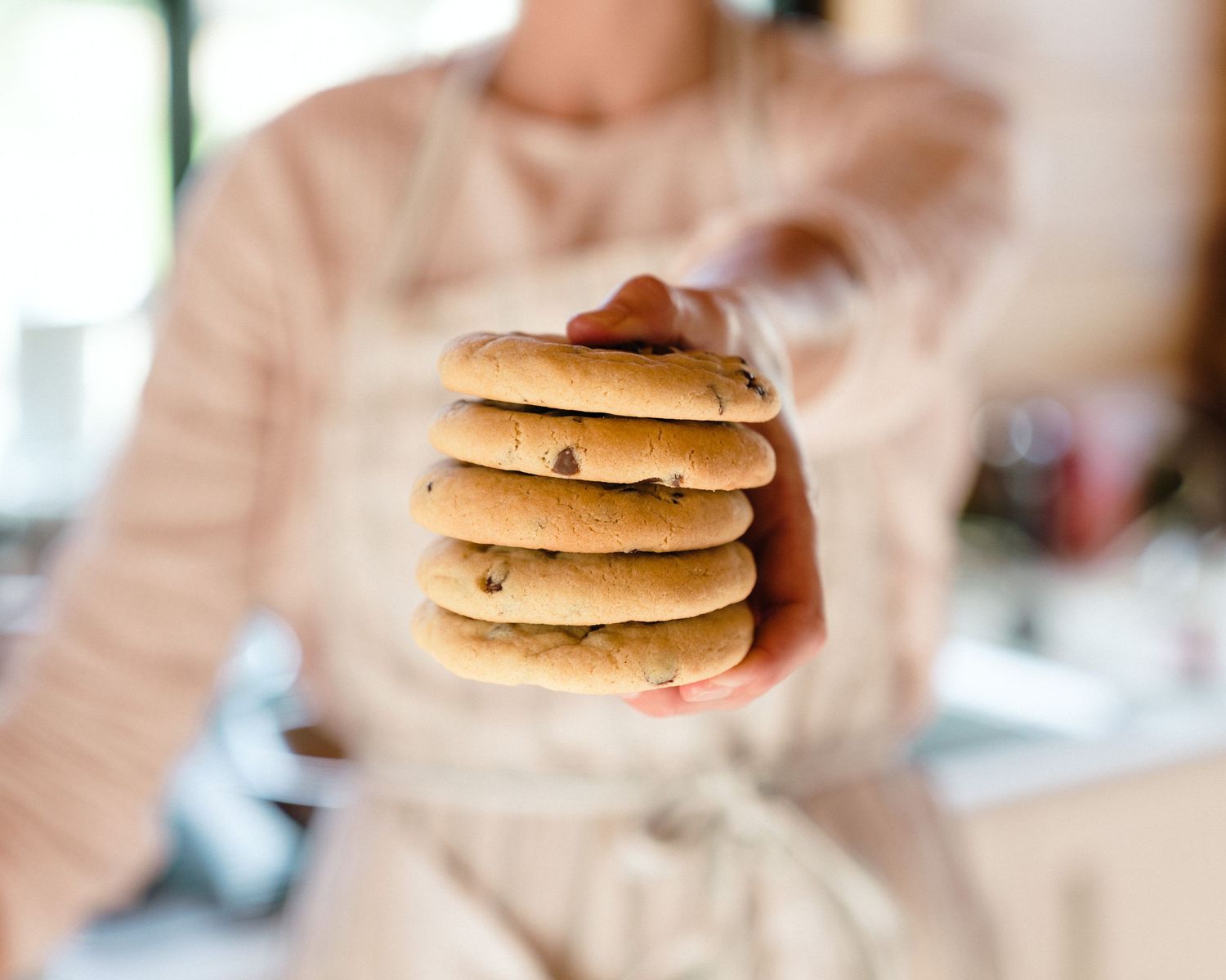 Someone holding a stack of freshly baked chocolate chip cookies against a blurred background.
