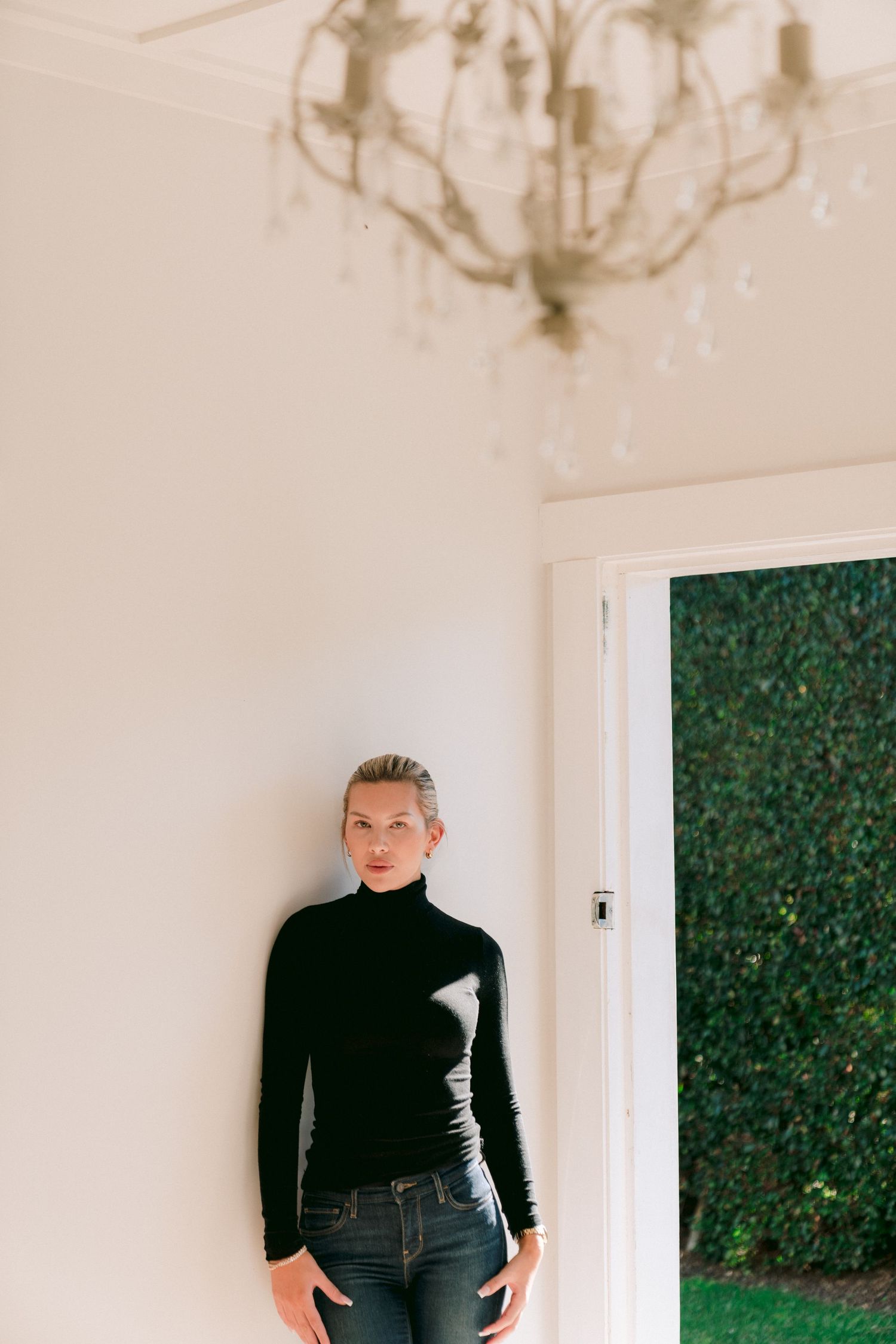 A series of minimalist fashion poses in black turtleneck and jeans against a white wall with greenery backdrop.