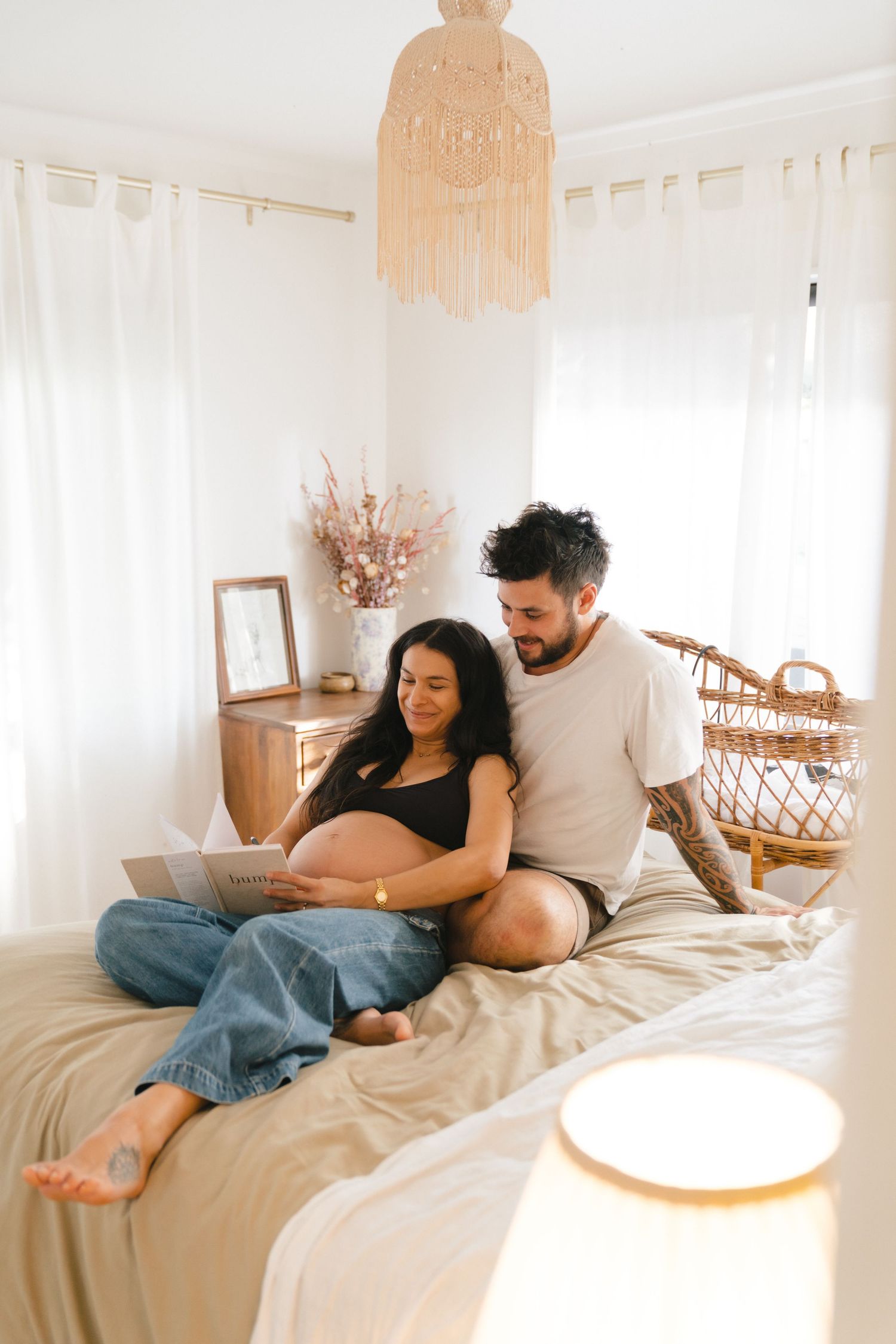 A couple relaxes together on a bed with a boho macrame chandelier and white curtains in a bright sunlit bedroom.