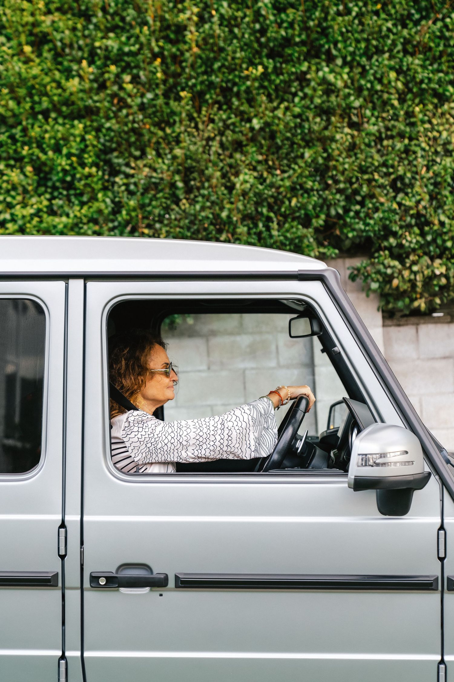 Driver in striped outfit at the wheel of a silver Mercedes G-wagon with sunglasses and side mirror visible.
