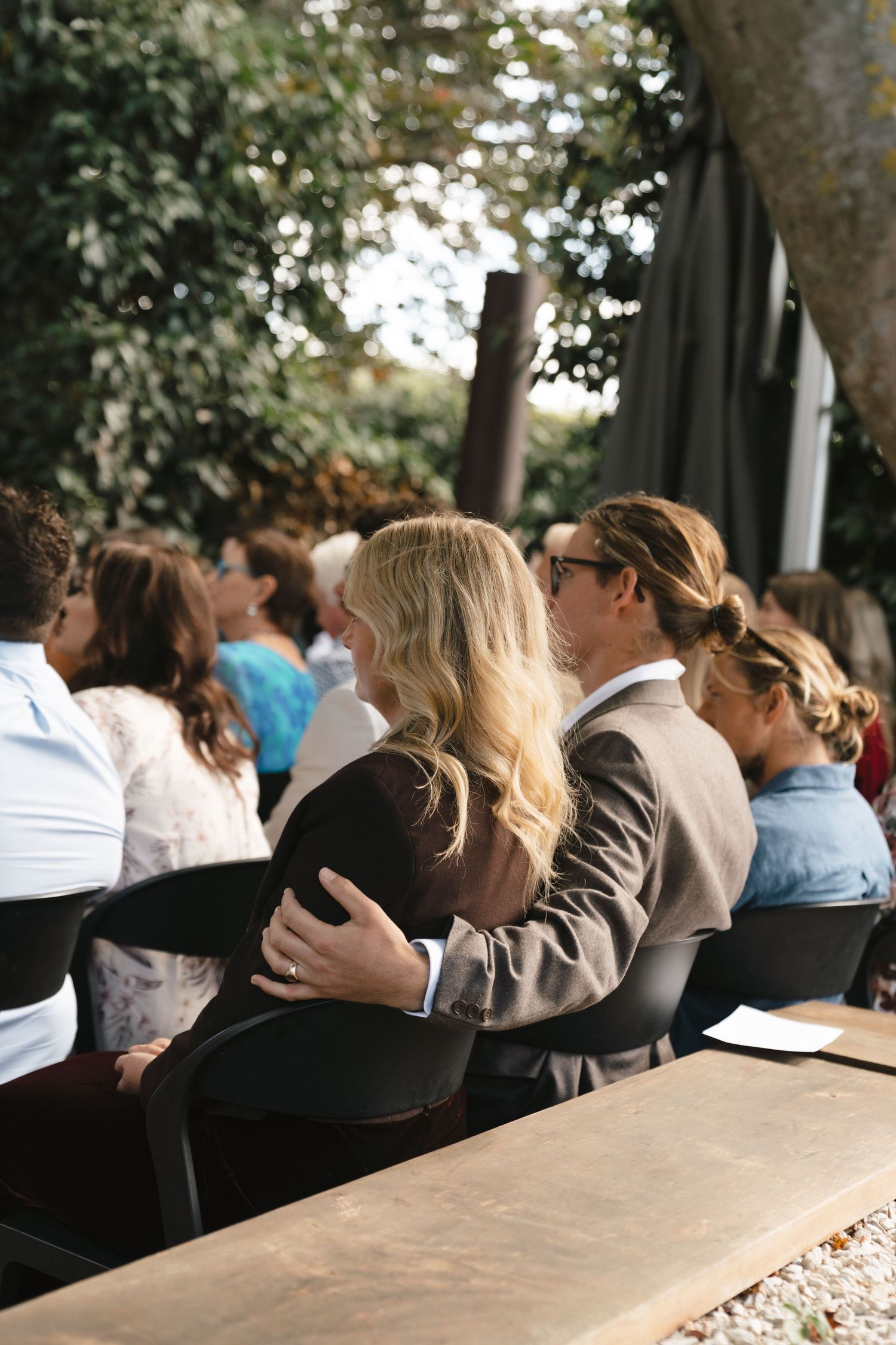 Wedding guests seated at outdoor ceremony venue under tree canopy.
