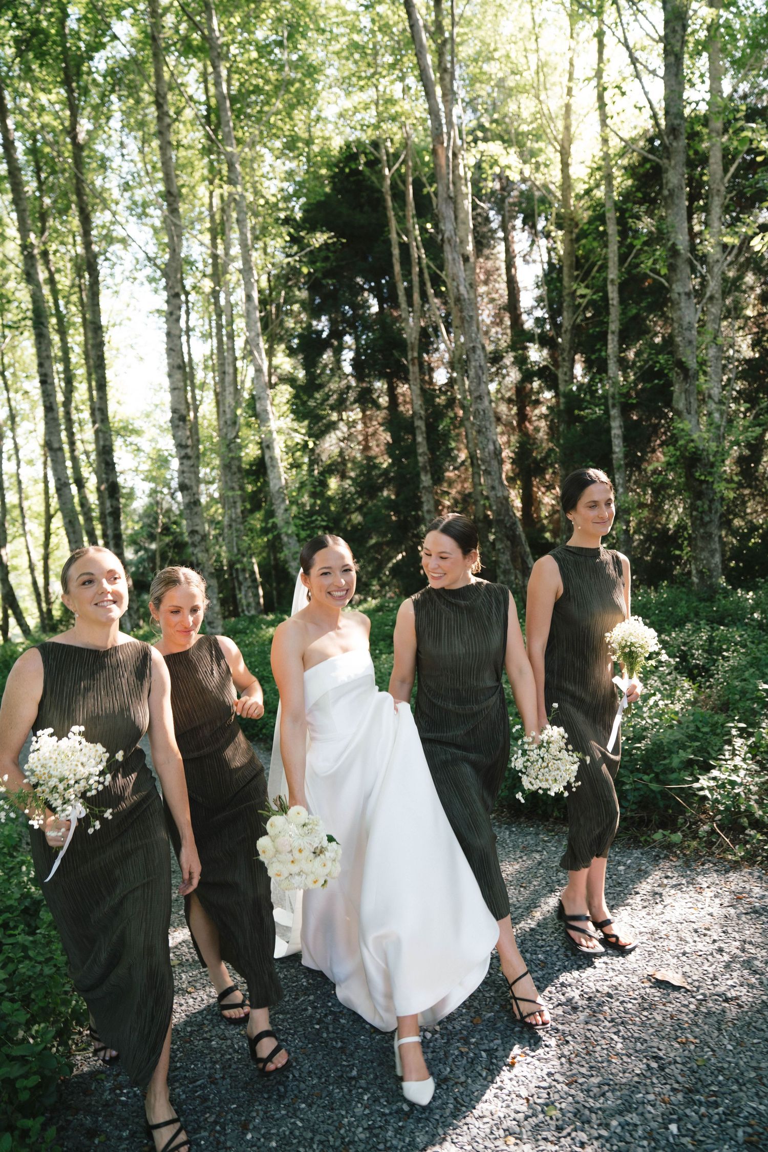 Wedding party in black dresses and white gown pose together on a tree-lined path in natural sunlight.