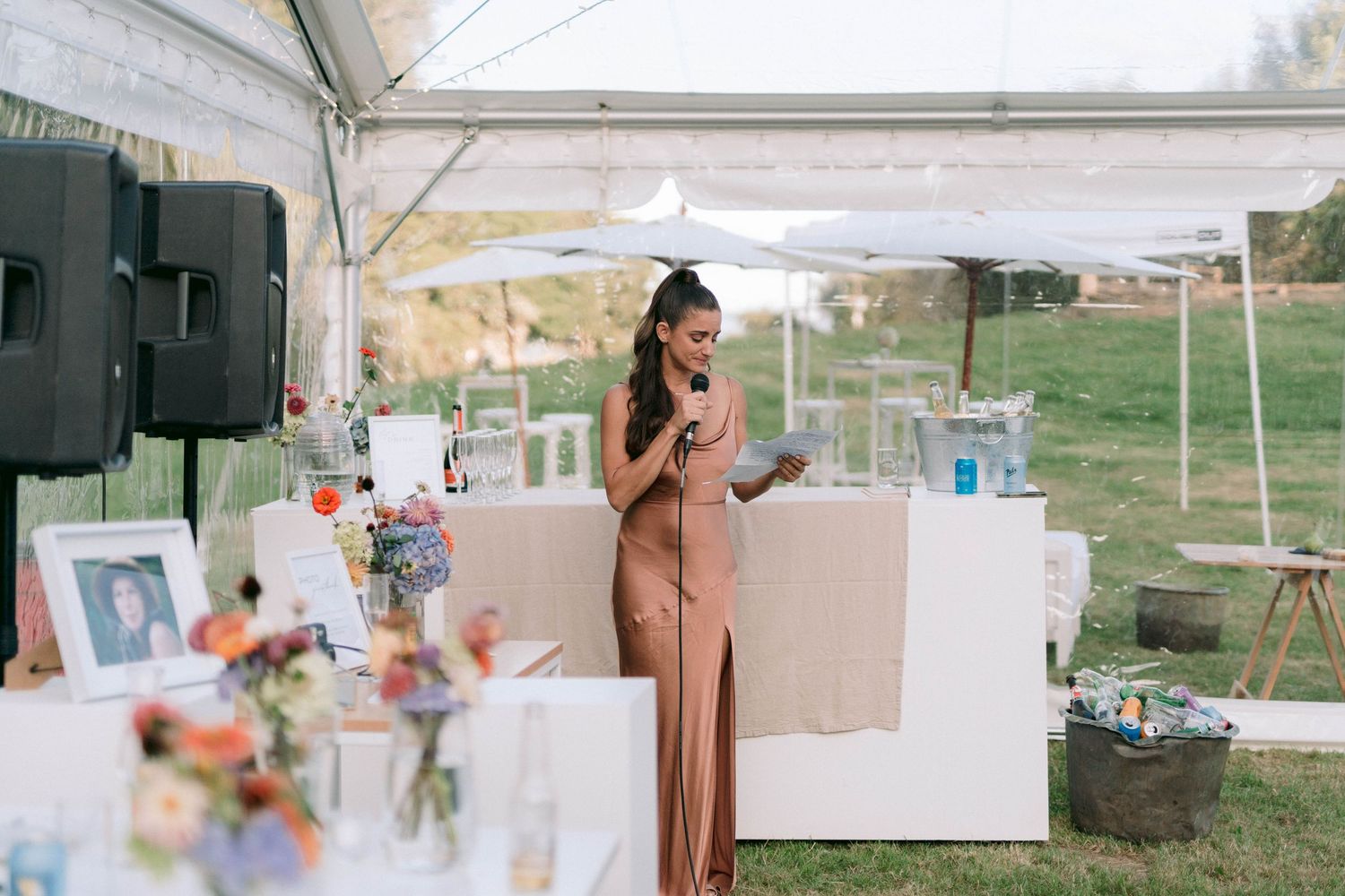 A woman in brown dress delivers a speech at an outdoor wedding reception.