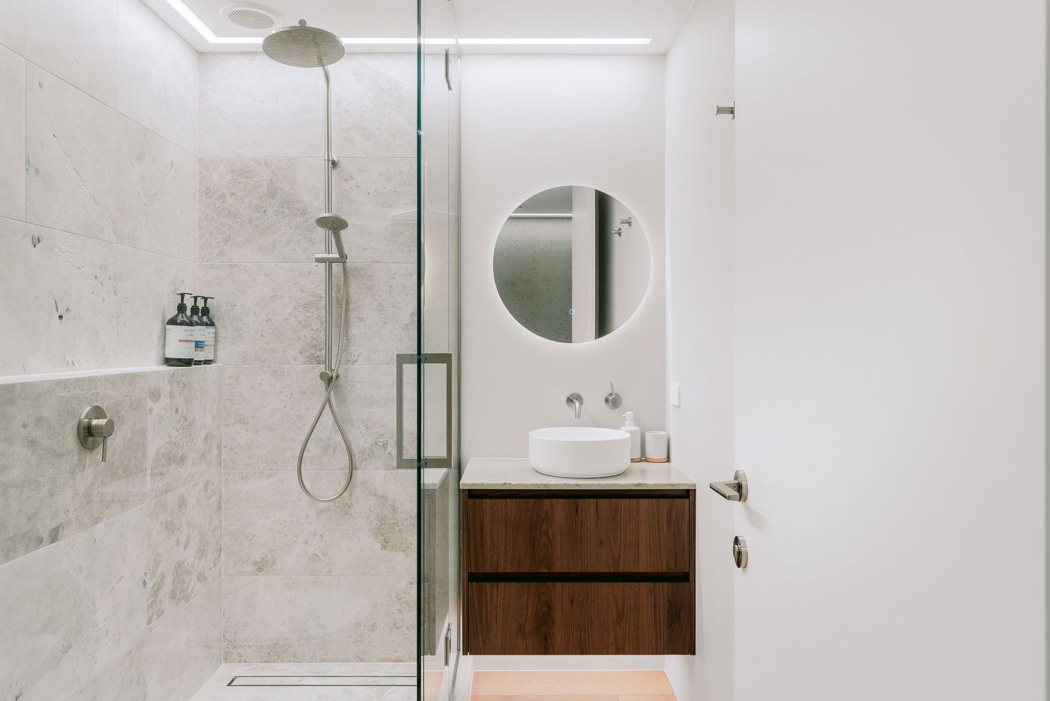 Sleek bathroom featuring marble walls, glass shower enclosure, and round mirror above wooden vanity.